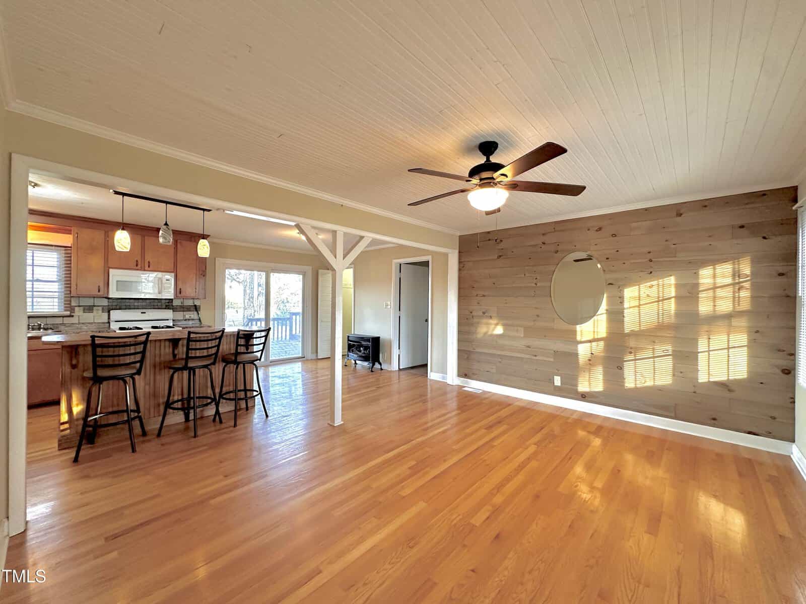 Open-concept living area with wood flooring, a ceiling fan, and a partial wood-paneled accent wall featuring a round mirror. The space transitions into a kitchen with a breakfast bar and pendant lighting. A large window offers natural light.
