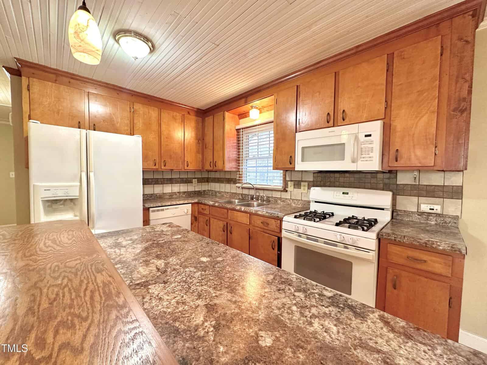 A kitchen with wooden cabinets, a patterned countertop, and tile backsplash. Features include a white refrigerator, microwave, and stove. Two pendant lights hang from the white paneled ceiling above the counter.