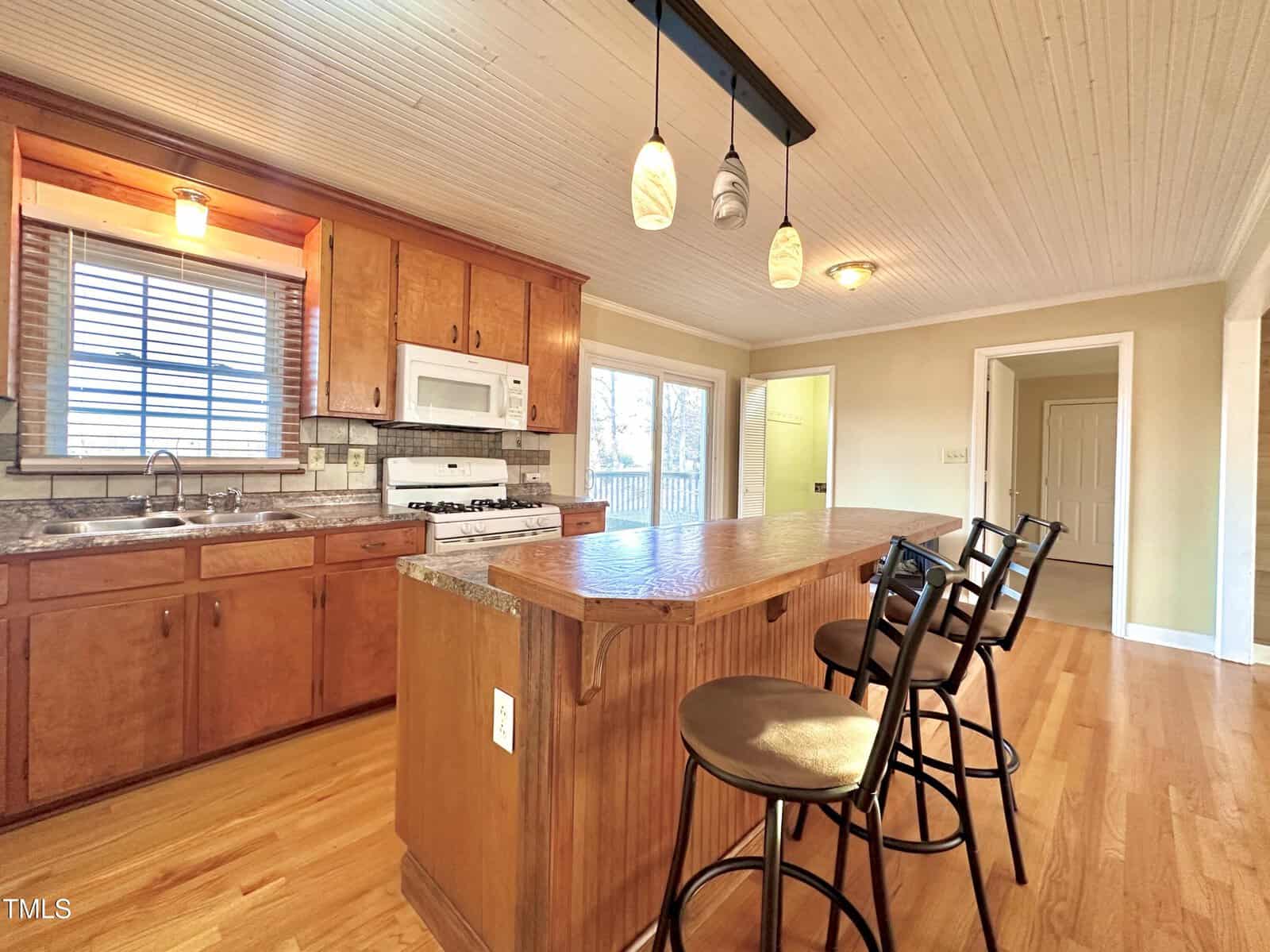A warm, cozy kitchen with wooden cabinets and flooring. It features a kitchen island with granite countertops and three bar stools. There is a gas stove, microwave, and a window above the sink letting in natural light.