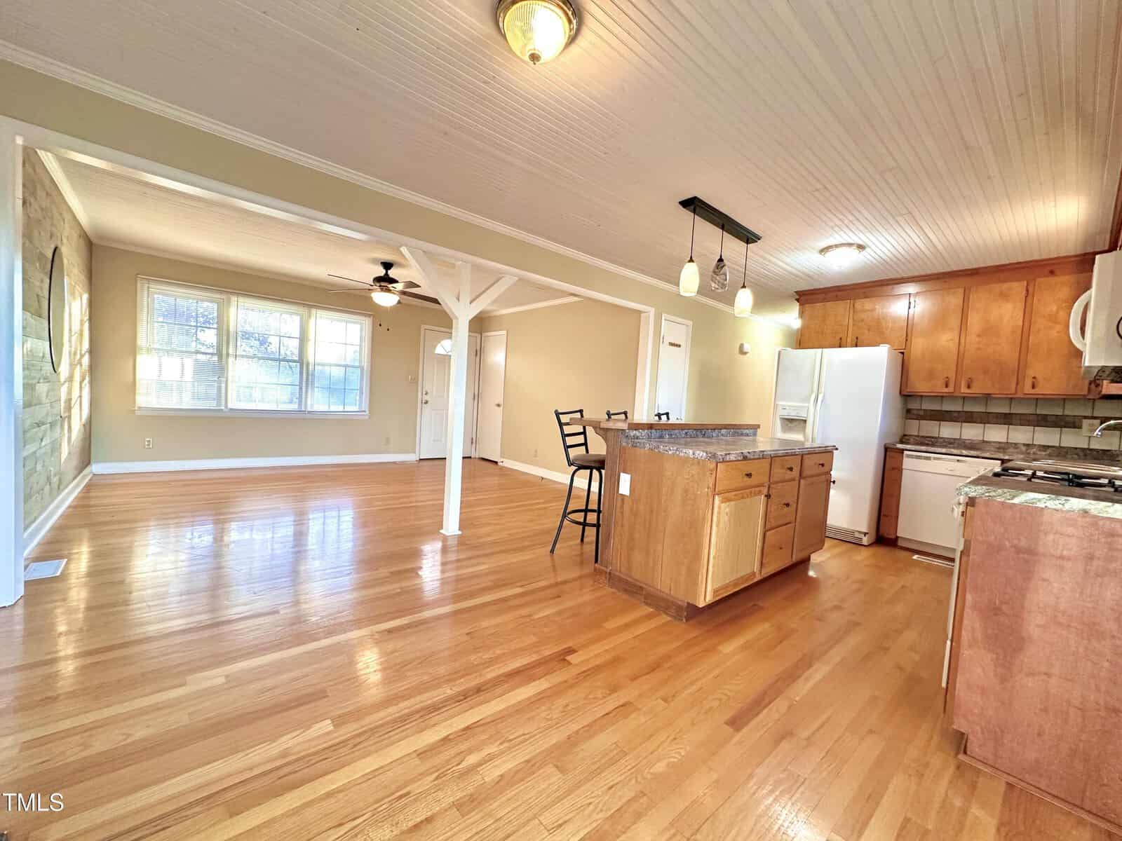 Spacious open-plan interior with hardwood floors, featuring a kitchen with wooden cabinets and a central island, pendant lighting above. Adjacent is a living area with large windows allowing natural light and a ceiling fan. Neutral colors throughout.