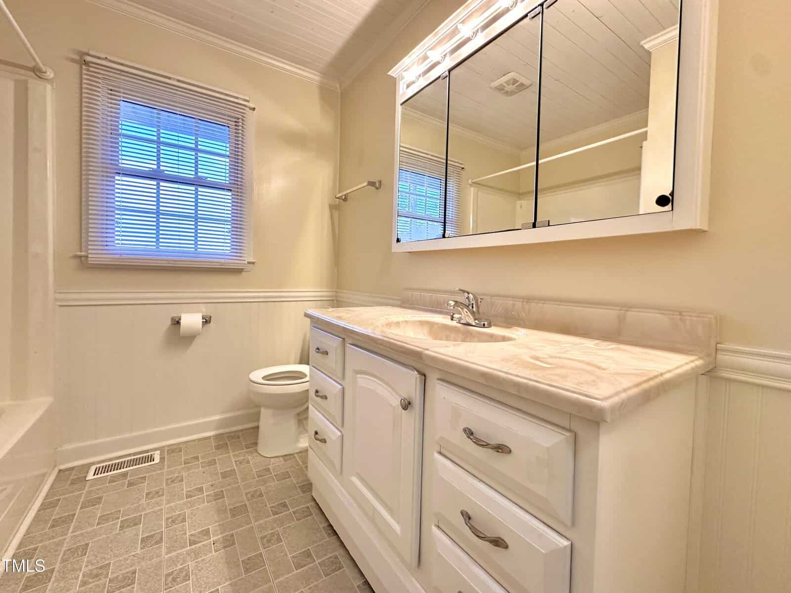 A bathroom features light beige walls, a large mirror above a white sink cabinet, and a toilet next to it. The floor is tiled in a gray pattern, and a window with blinds allows natural light in. The shower is partially visible on the left.
