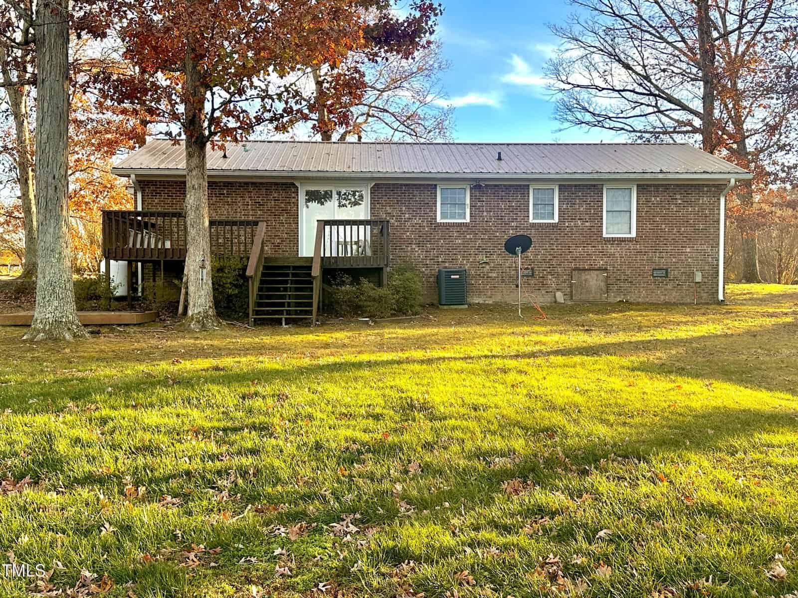 A single-story brick house with a metal roof is surrounded by autumn trees. A small wooden deck with stairs leads to the back door. A satellite dish is nearby on a grassy lawn, with sunlight filtering through the leaves.