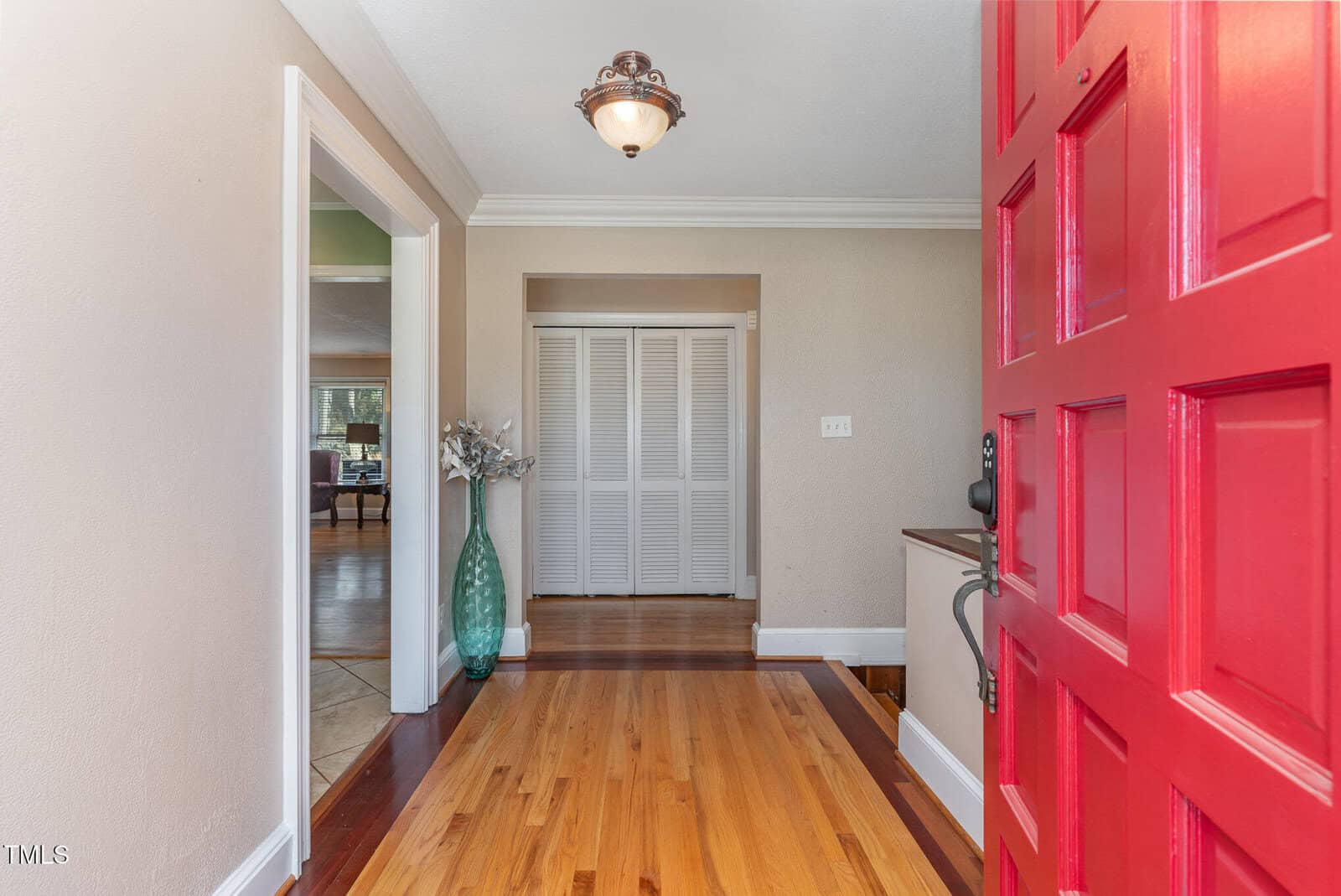 Open red door reveals an entryway with wood floor, beige walls, a ceiling light, and a large glass vase with white flowers. White double doors are straight ahead, leading into another room on the left.