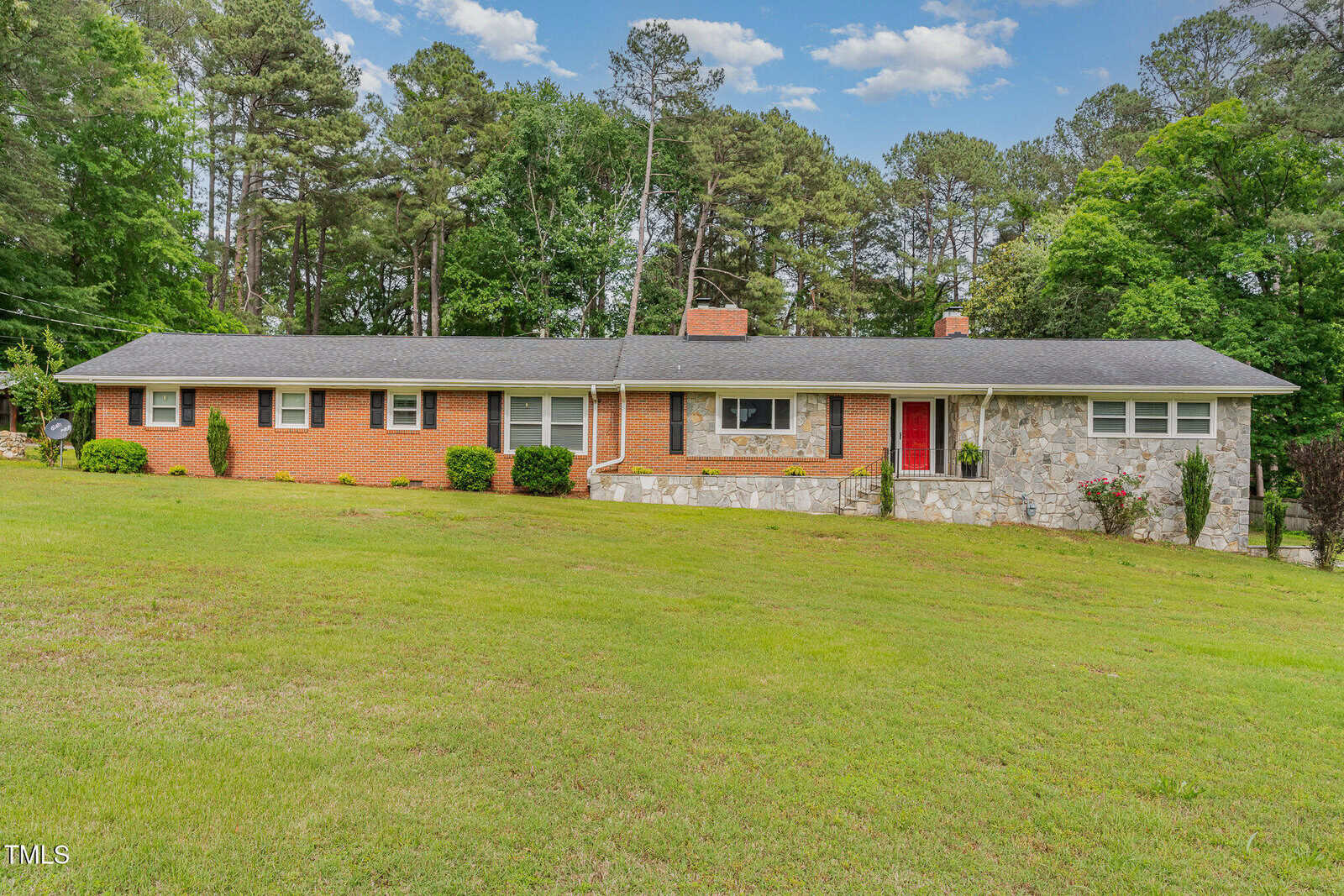 A single-story ranch-style house with a brick and stone exterior, black shingle roof, red front door, and a large grassy yard. Trees and shrubs surround the property, with a forested area in the background.