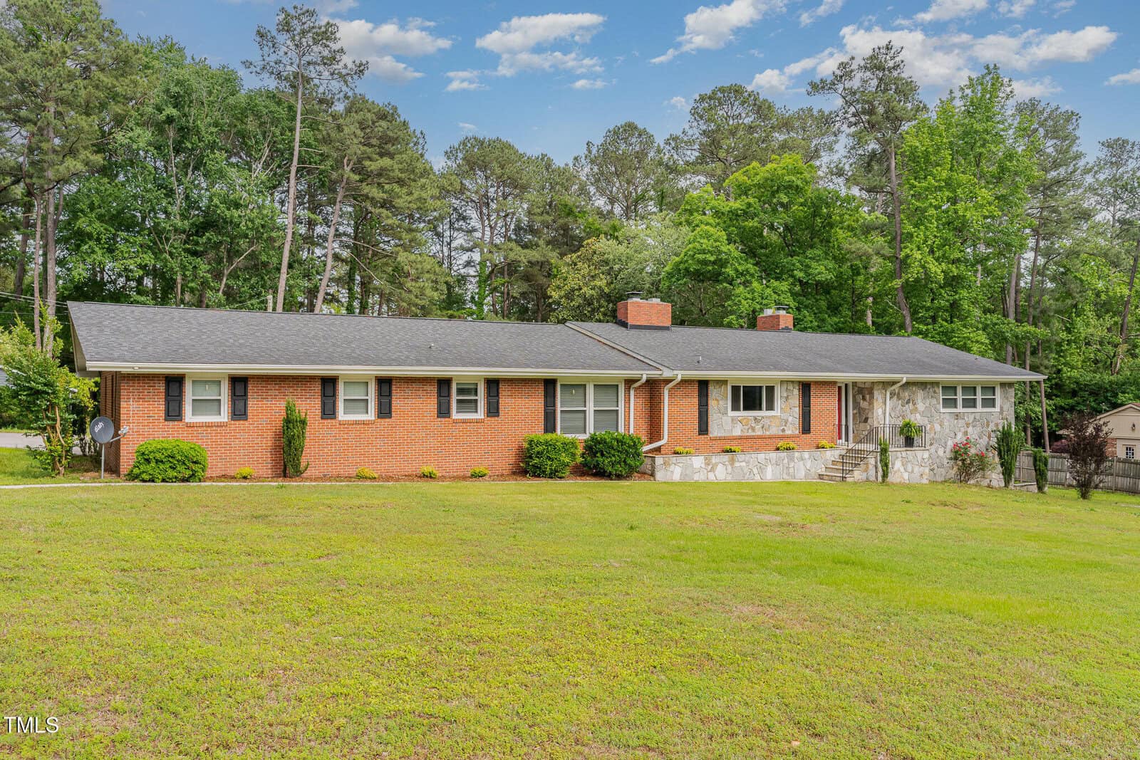 Single-story red brick house with a dark roof, white trim, large front lawn, stone steps, and surrounded by tall green trees under a partly cloudy blue sky.