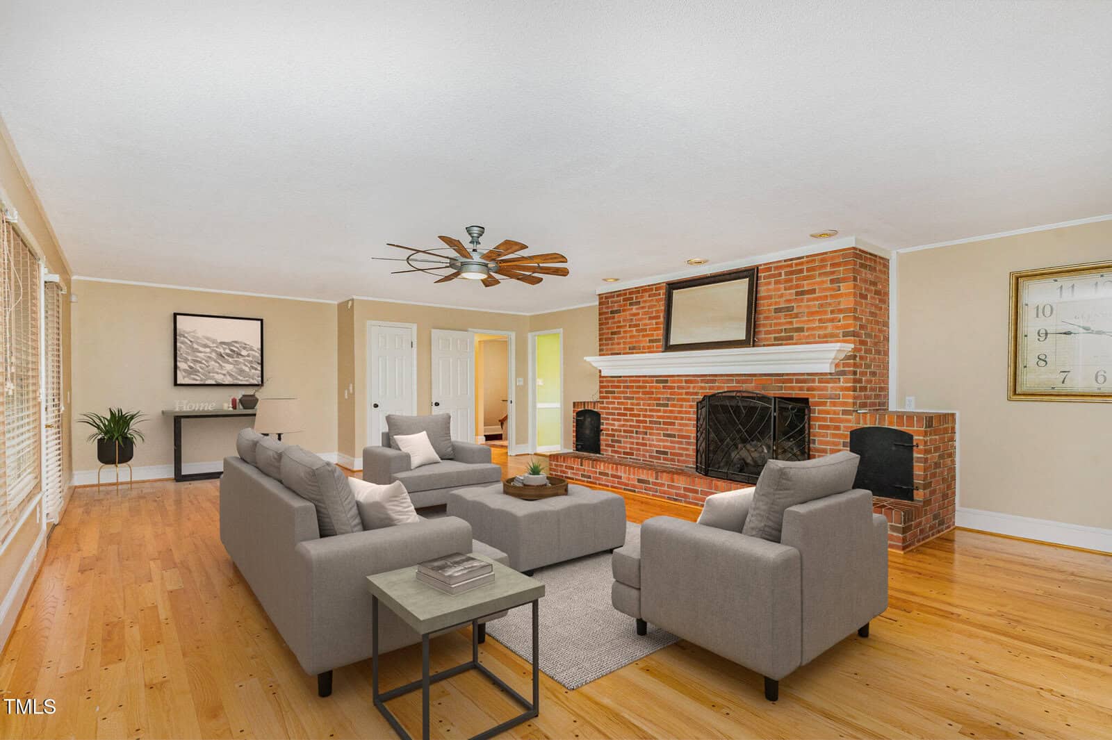 Bright living room with light wood floors, gray sofas, a brick fireplace, a large wall clock, and a ceiling fan. Windows line one wall, and a potted plant sits in the corner near a framed picture.