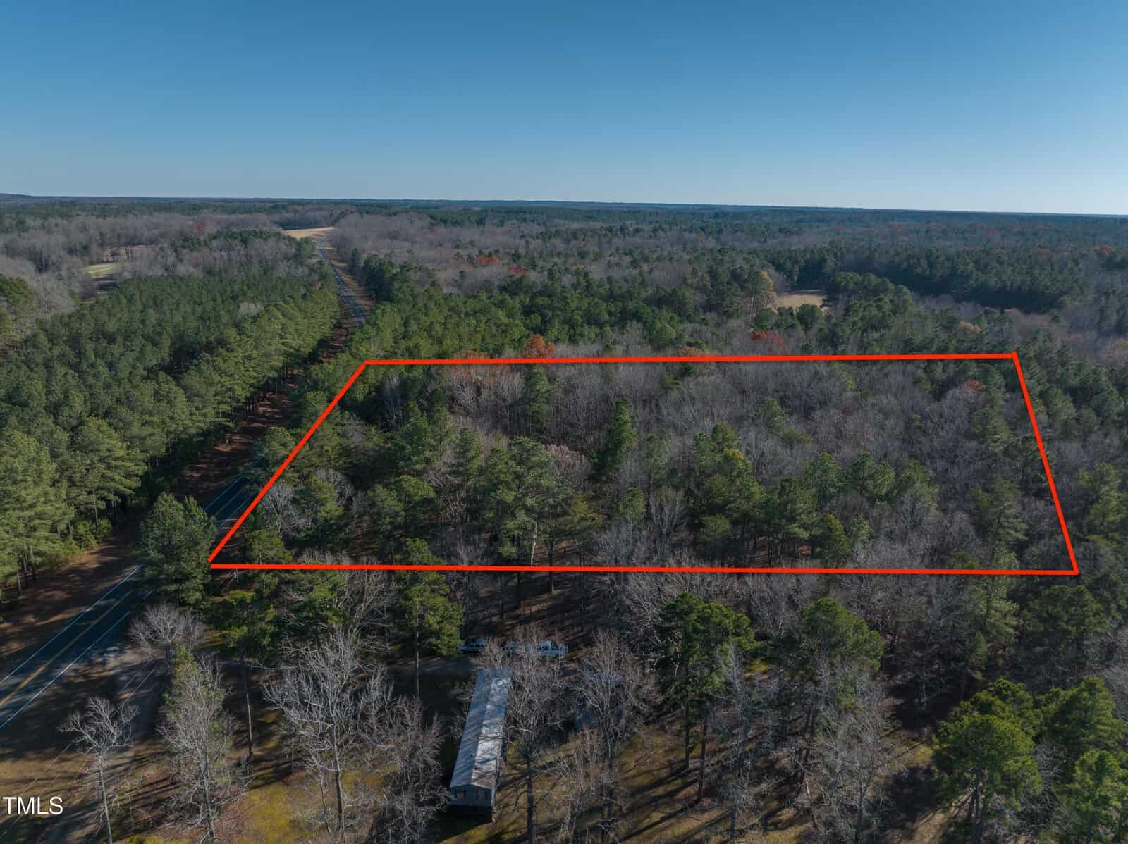 Aerial view of a forested area bordered by a red rectangle. Adjacent to the forest is a road on the left and a small building at the bottom. The sky is clear and blue.