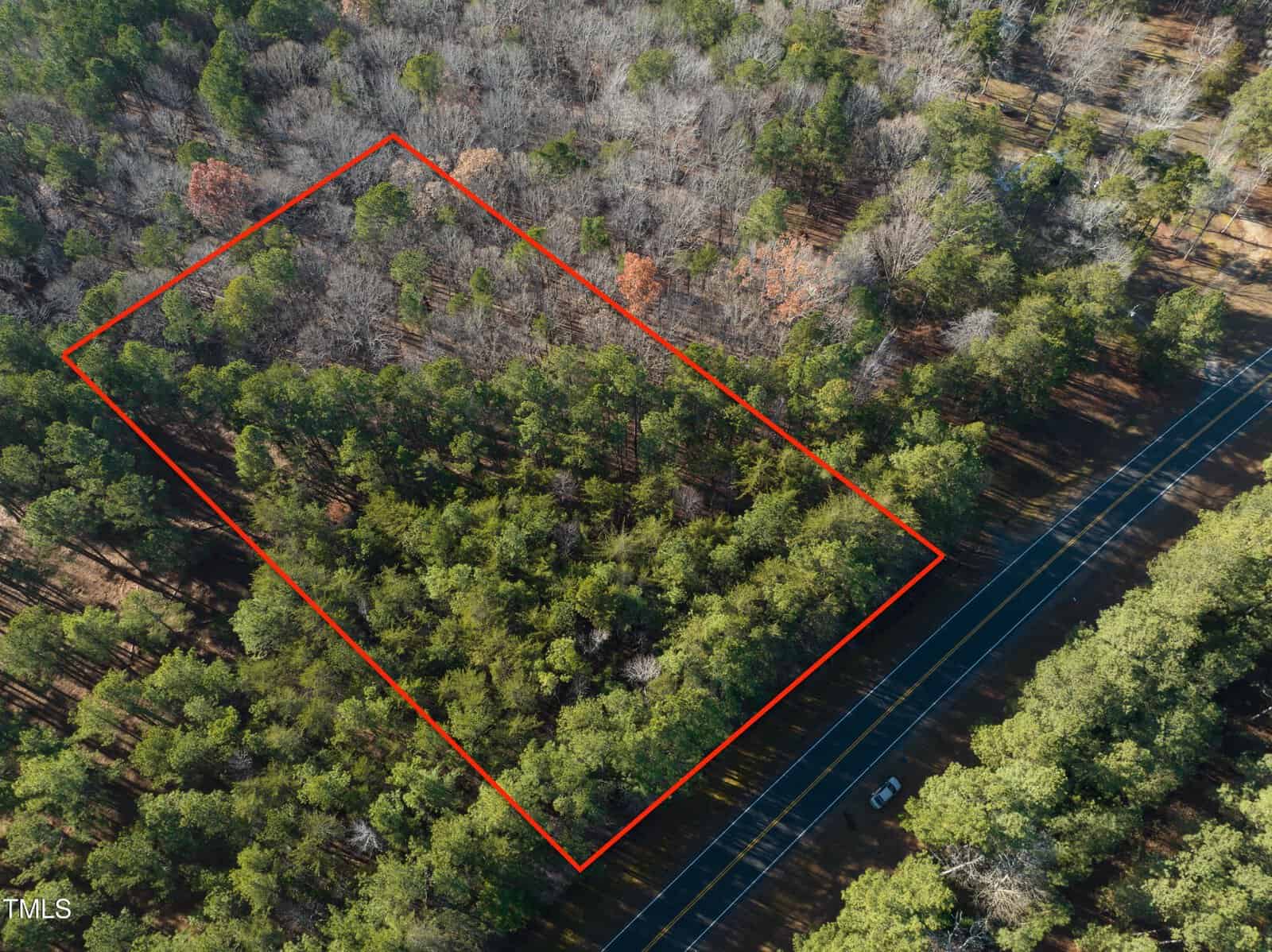 Aerial view of a rectangular plot of forested land outlined in red. The area is densely covered with trees. A road with a vehicle is visible on the lower right side, adjacent to the plot.