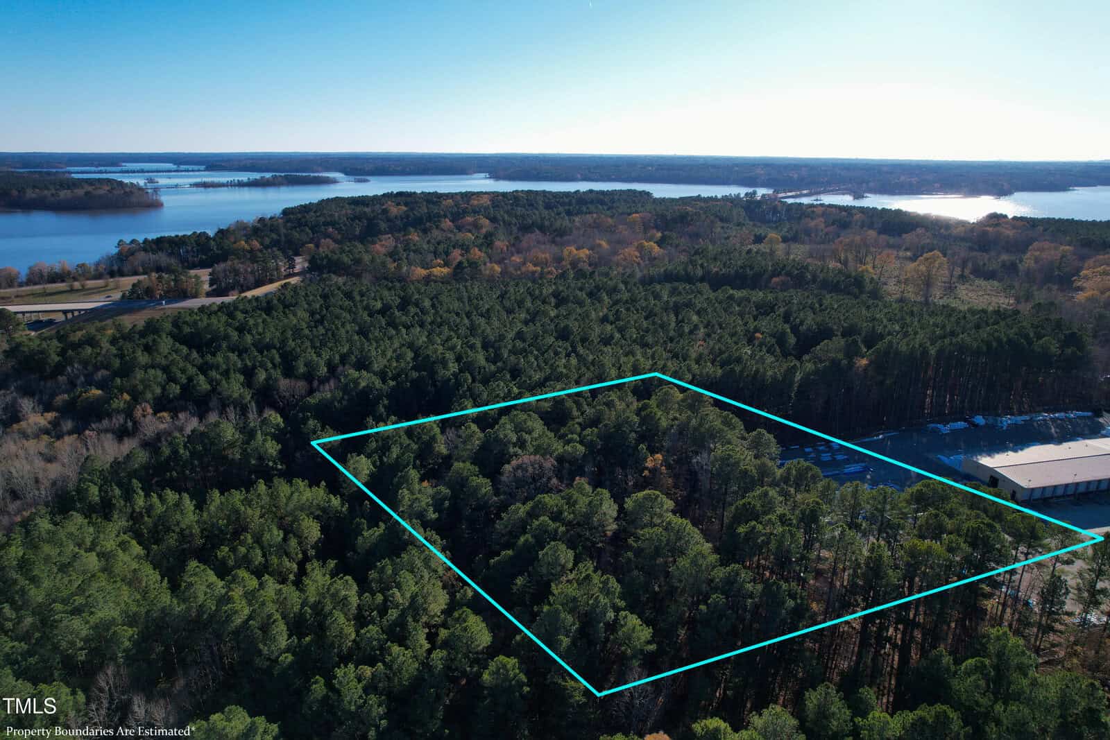 Aerial view of a forested area with a rectangular plot outlined in blue. Beyond the trees, a large body of water is visible. A building is partially visible on the right side. The sky is clear and blue.