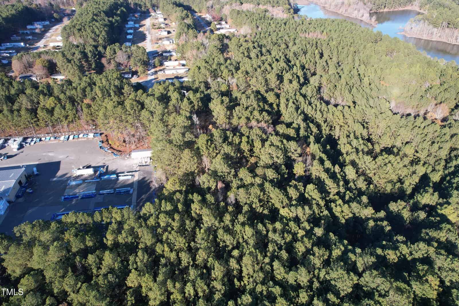Aerial view of a densely wooded area with several buildings and trailers at the bottom left. A winding road runs through the trees, and a body of water is visible in the upper right corner. The landscape is predominantly forested.