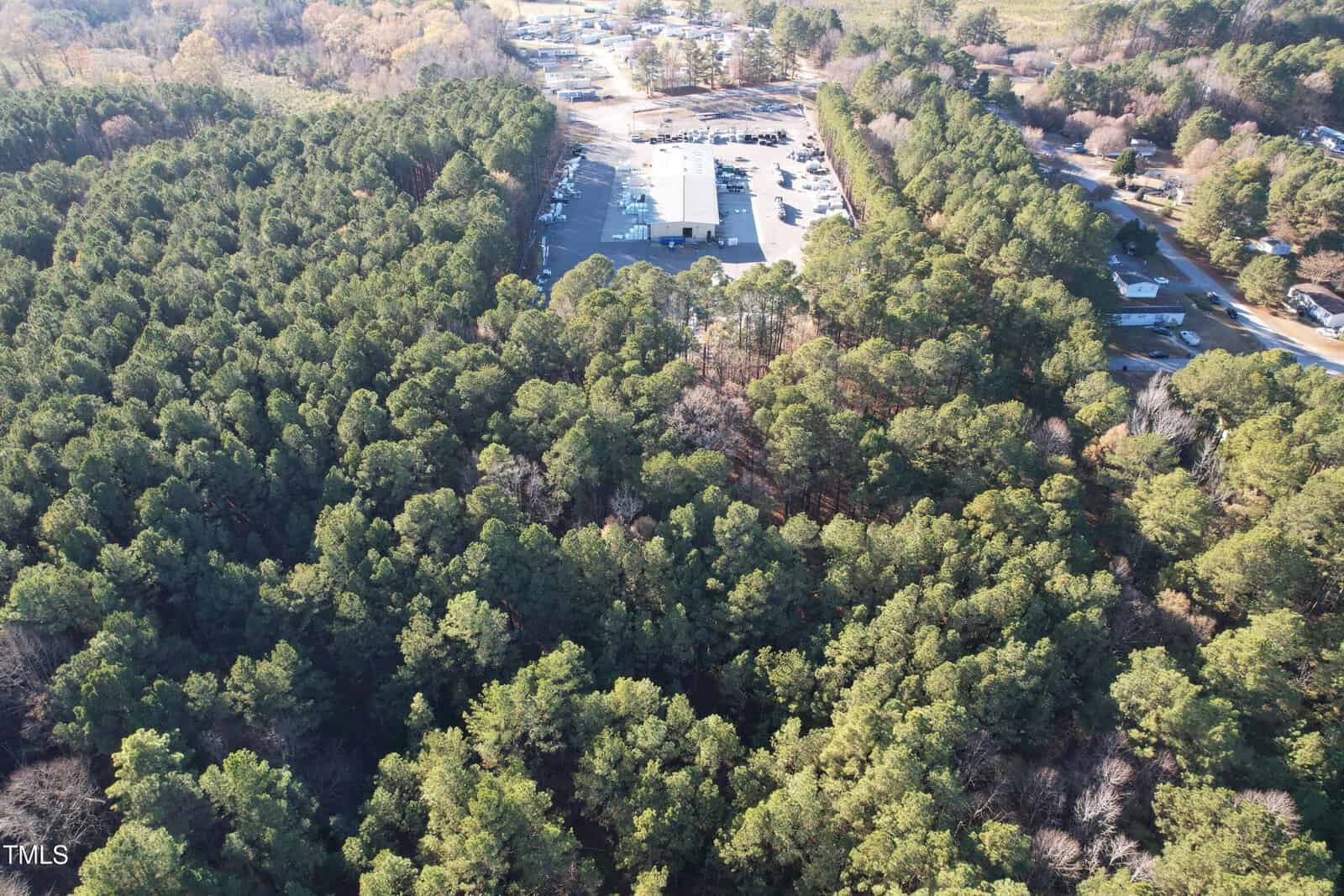 Aerial view of a forested area with dense trees surrounding an industrial facility and a parking lot. Roads are visible on the right and top sides, with a mix of trees and open spaces scattered throughout the landscape.