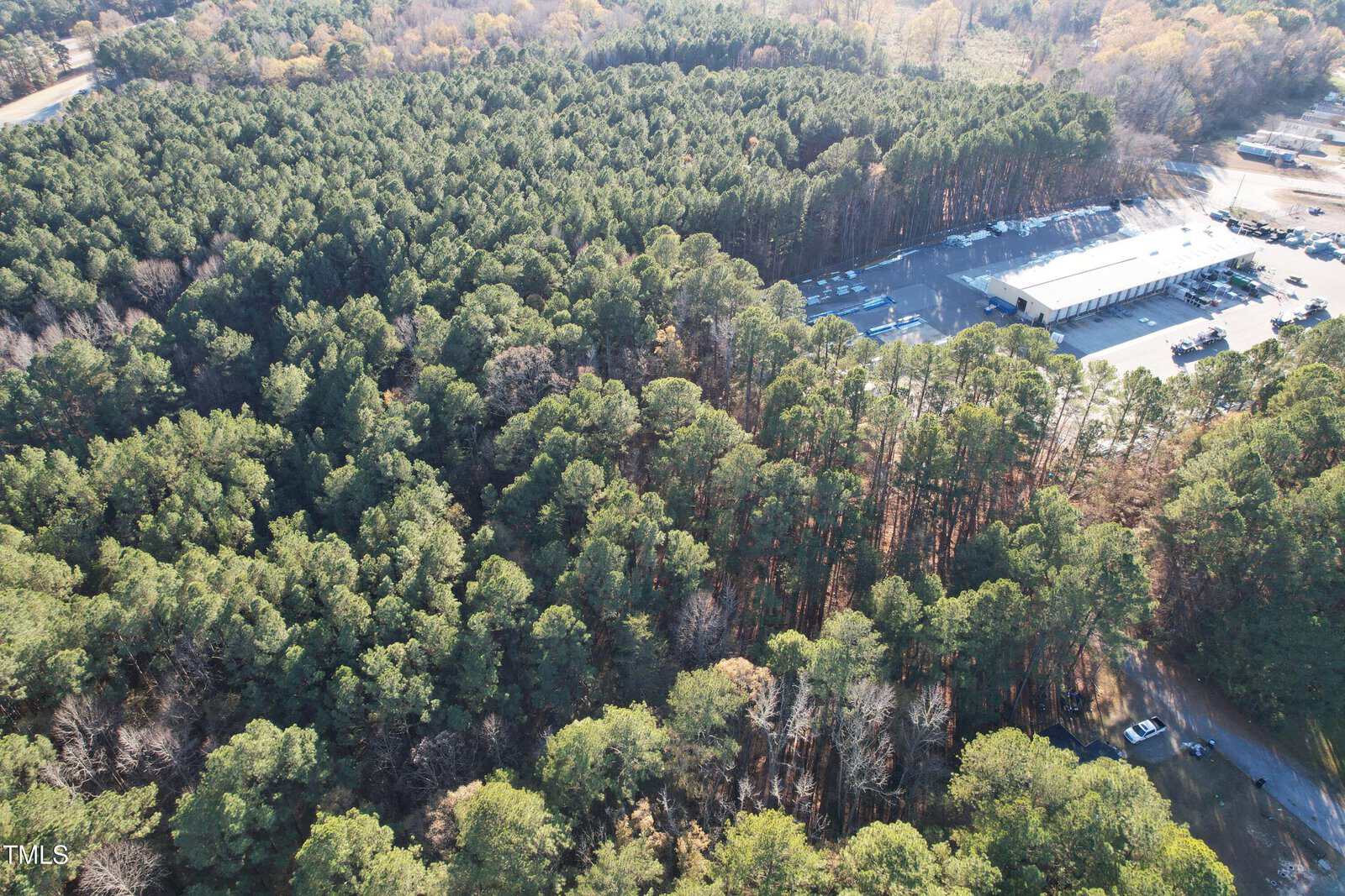 Aerial view of a dense forest with a large building complex in the top-right corner. The scene includes surrounding roads and scattered smaller structures, with trees showing a mix of green and autumn hues.