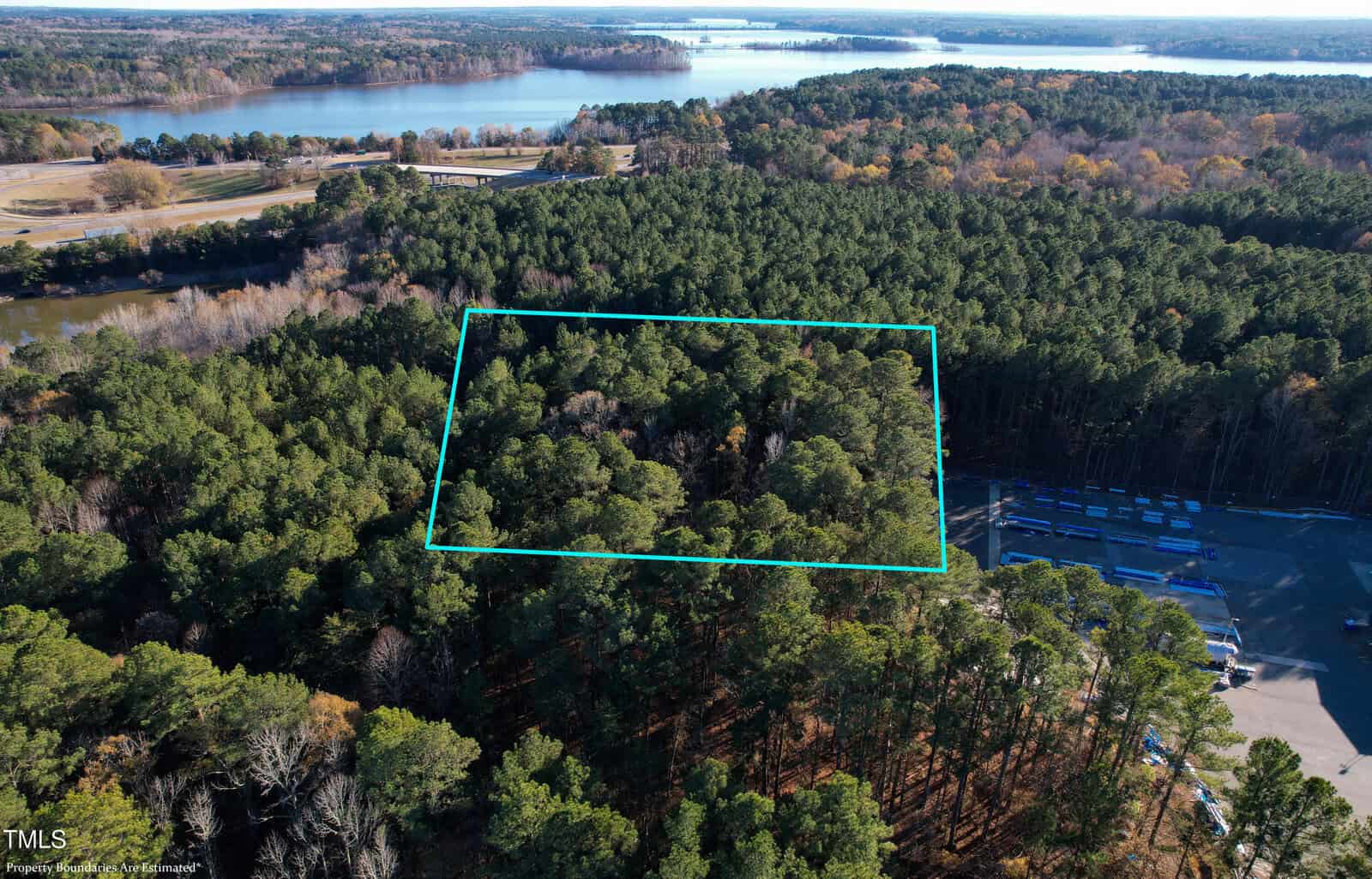 Aerial view of a dense forested area bordered by a teal outline, with a body of water visible in the background. A structure with several parked vehicles is located near the trees. The landscape includes a mix of forest, water, and open land.
