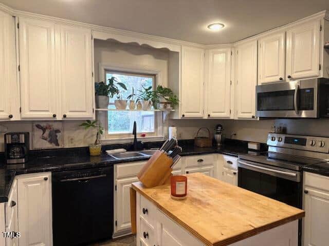 A modern kitchen features white cabinets and black countertops. A wood-topped island stands in the center, with a knife block and candle on it. Appliances include a stainless steel oven, microwave, dishwasher, and a coffee maker. A window has potted plants.