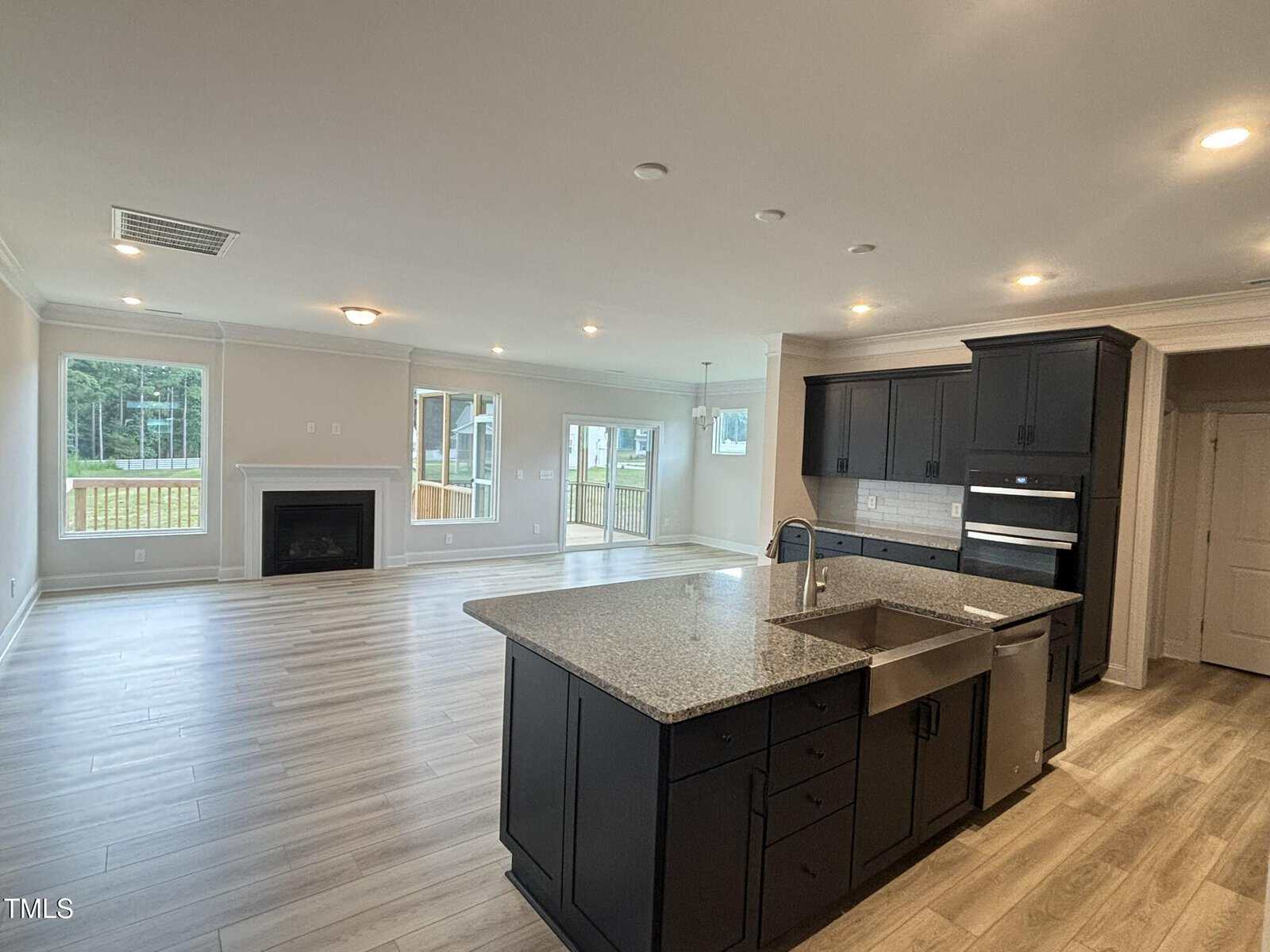 Spacious open-concept kitchen and living area featuring dark cabinets, a large island with a sink, built-in appliances, light wood flooring, fireplace, and large windows overlooking an outdoor deck.
