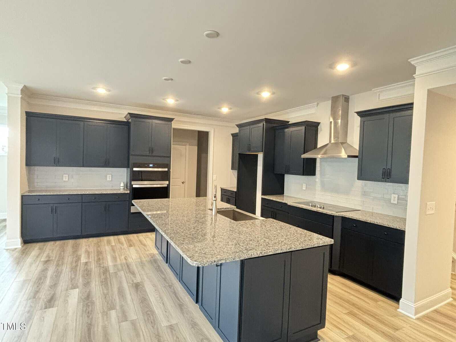 Modern kitchen with dark cabinets, stainless steel appliances, a large granite island with a sink, light wood flooring, and recessed ceiling lights. The space is clean and uncluttered.
