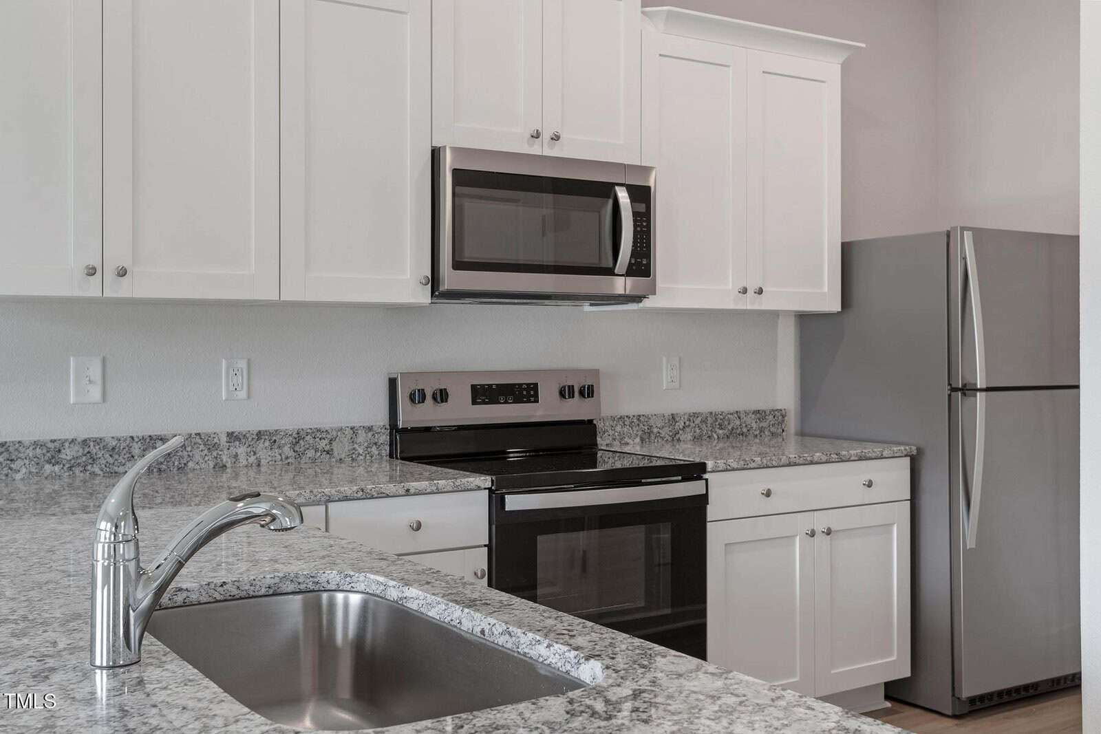 Modern kitchen with white cabinets, a stainless steel refrigerator, oven, and microwave. Granite countertops, a sink with a chrome faucet, and tiled backsplash are visible. Bright and minimalist design.