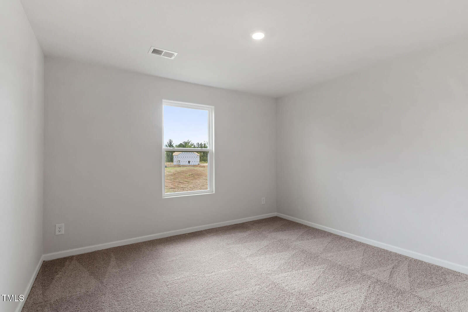 An empty room with light gray walls and beige carpet. A single, small window is on one wall, showing a view of a grassy field and a white building outside. The ceiling has a recessed light fixture.