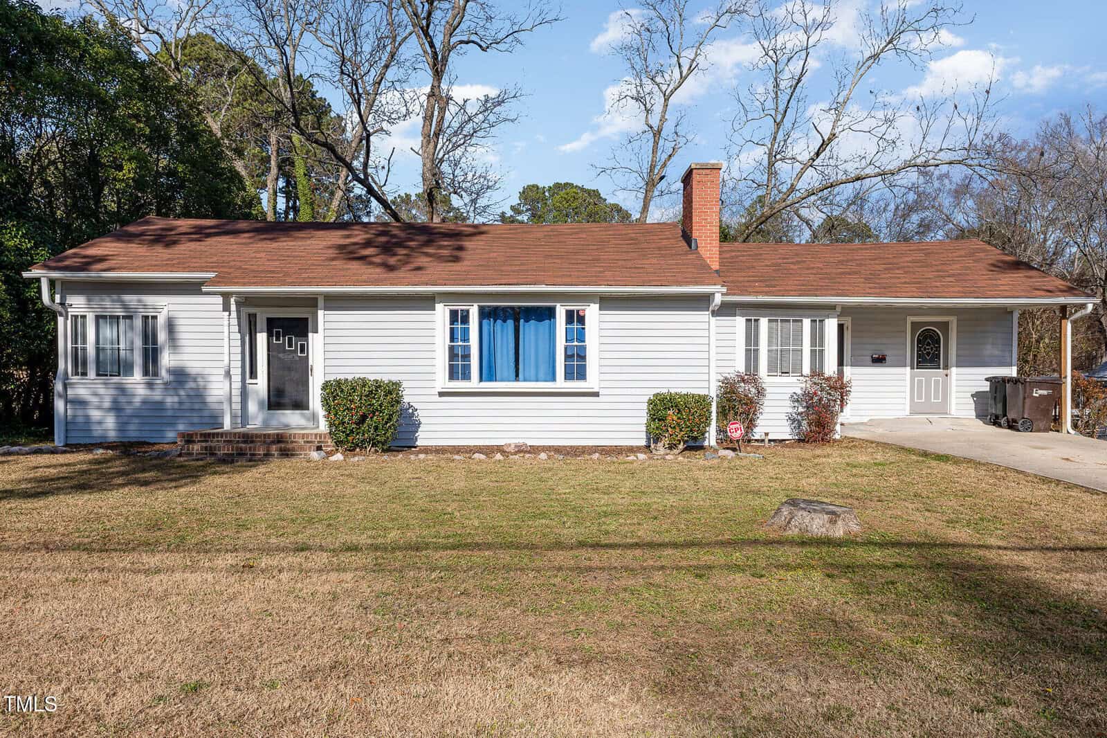 Single-story house with light gray siding, brown shingle roof, and blue curtains in the front window. There is a small front yard with sparse grass, shrubs by the house, and a driveway on the right side.