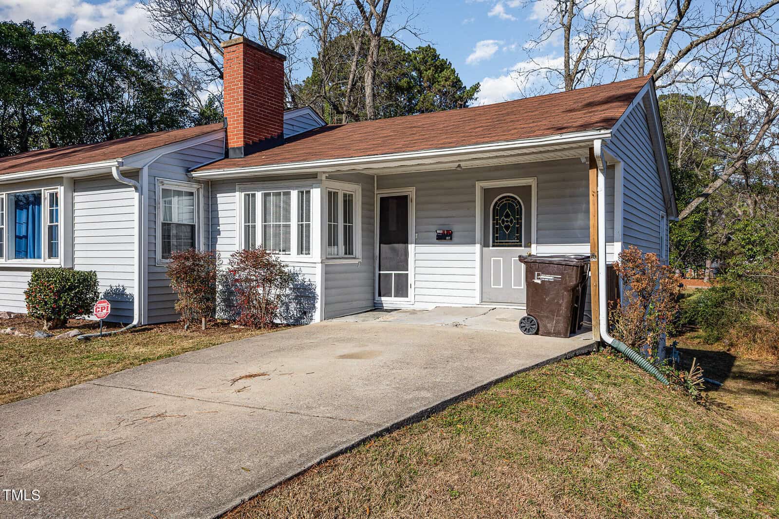 A small single-story gray house with a red roof, a brick chimney, a driveway, and a brown trash bin near the front door. There are a few bushes along the front and the lawn is mostly brown.