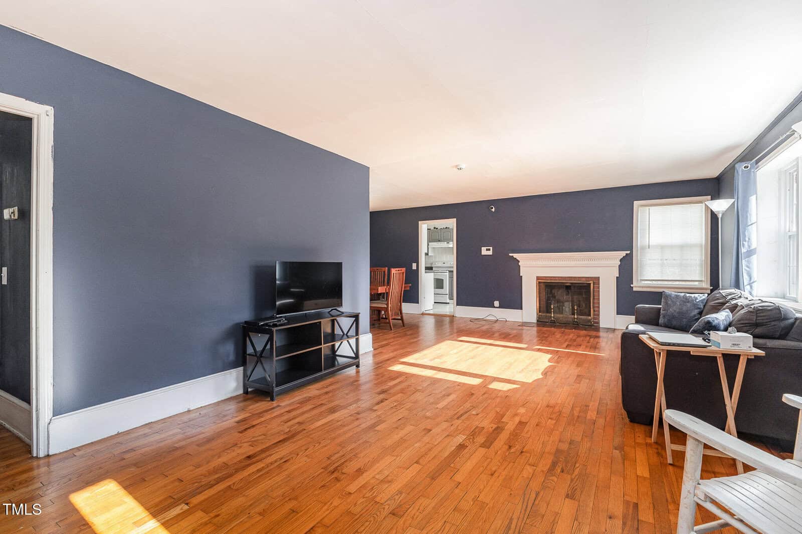 A living room with dark blue walls, hardwood floors, a couch, a TV on a stand, a white fireplace, a window letting in sunlight, and a small dining area visible through an open doorway.