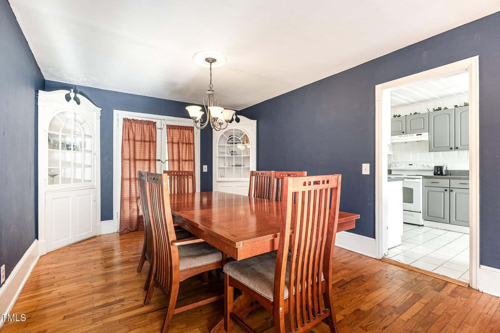 A dining room with a wooden table and six chairs, dark blue walls, hardwood floors, built-in white cabinets, a chandelier, and a view into a white kitchen.