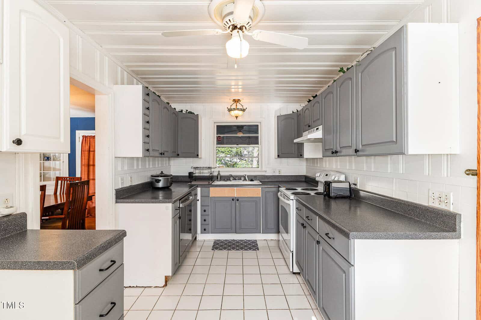 Bright kitchen with gray and white cabinets, white tile floor, black countertops, white appliances, a ceiling fan, and a window above the sink. Dining room is visible through the doorway on the left.