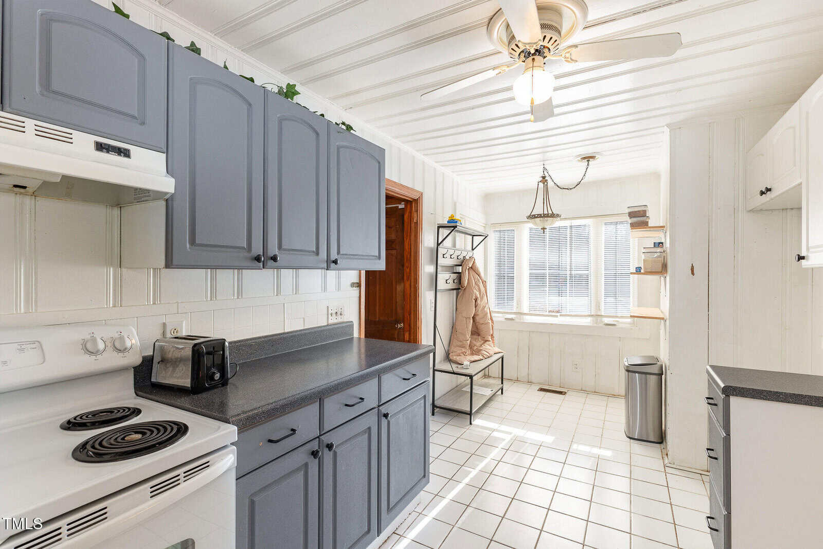 Bright kitchen with gray cabinets, white appliances, tile floor, and a ceiling fan. A bench with coat hooks and windowed breakfast nook are visible, along with a toaster, trash can, and overhead greenery accents.