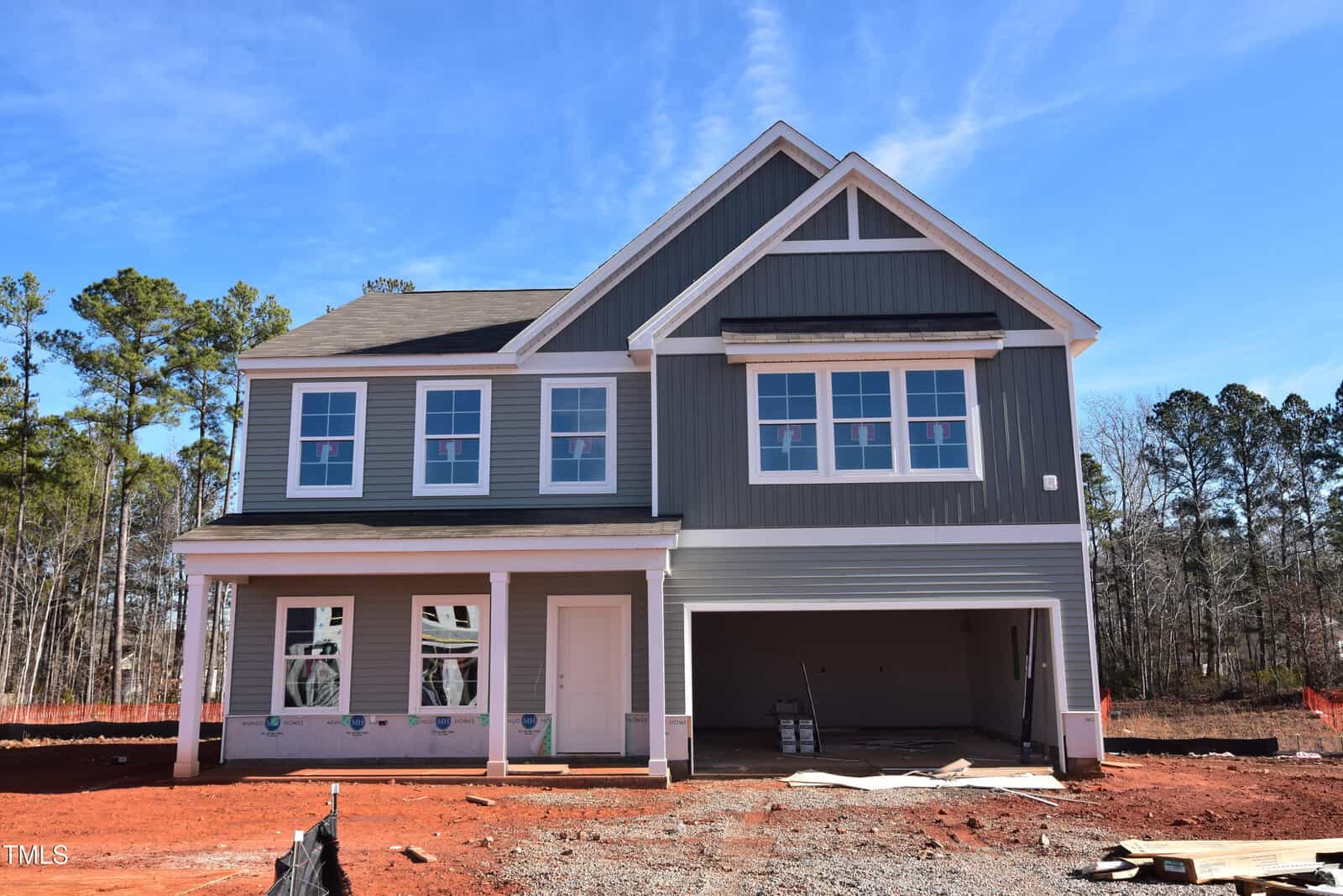 A two-story house under construction with gray siding, white trim, and multiple windows. The garage is open with building materials inside, and the yard is covered in red dirt. Trees are visible in the background under a blue sky.