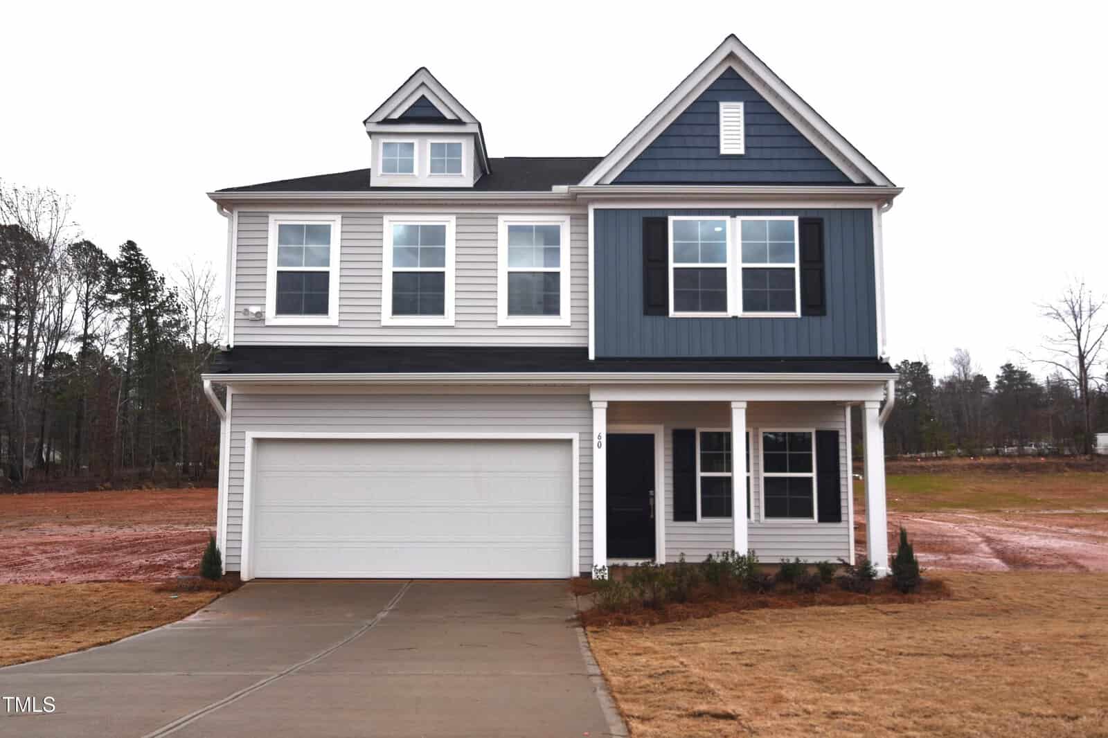 A two-story modern house with gray siding, white trim, and a blue accent section. There is a two-car garage, a covered front porch, and the yard appears to be unfinished with bare soil and minimal landscaping.