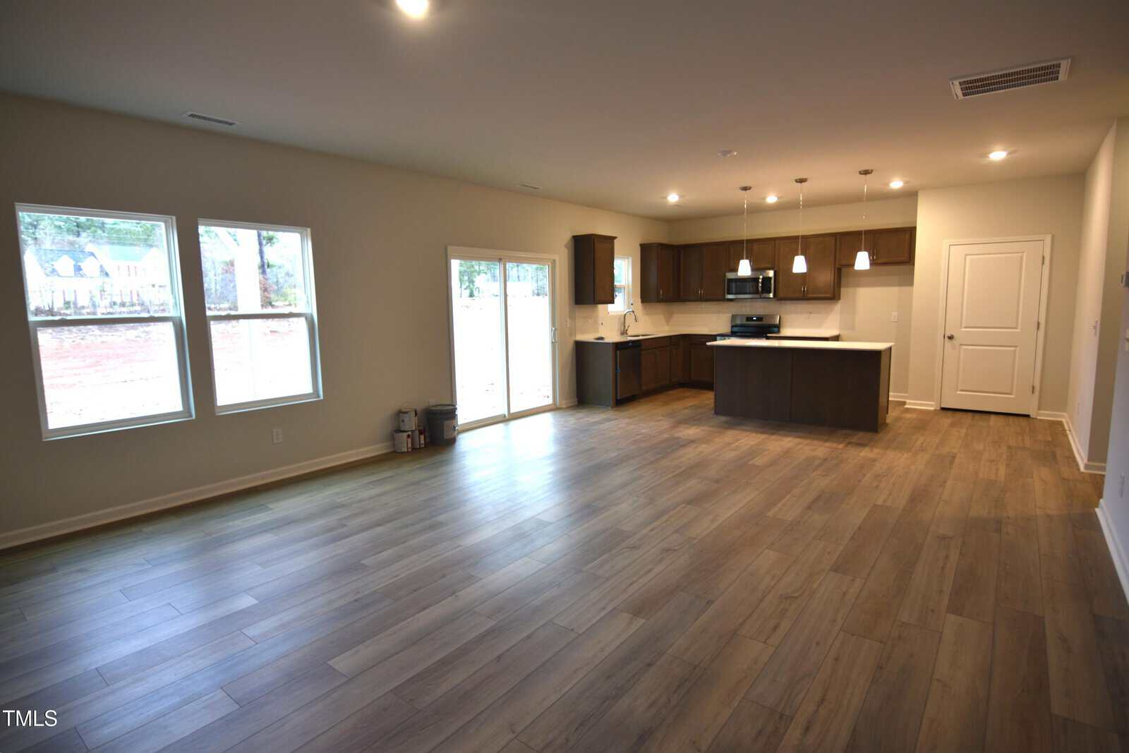 An empty open-plan living area with wood-look flooring, beige walls, three windows, a sliding glass door, and a kitchen with dark cabinets, stainless steel appliances, and pendant lights over a central island.