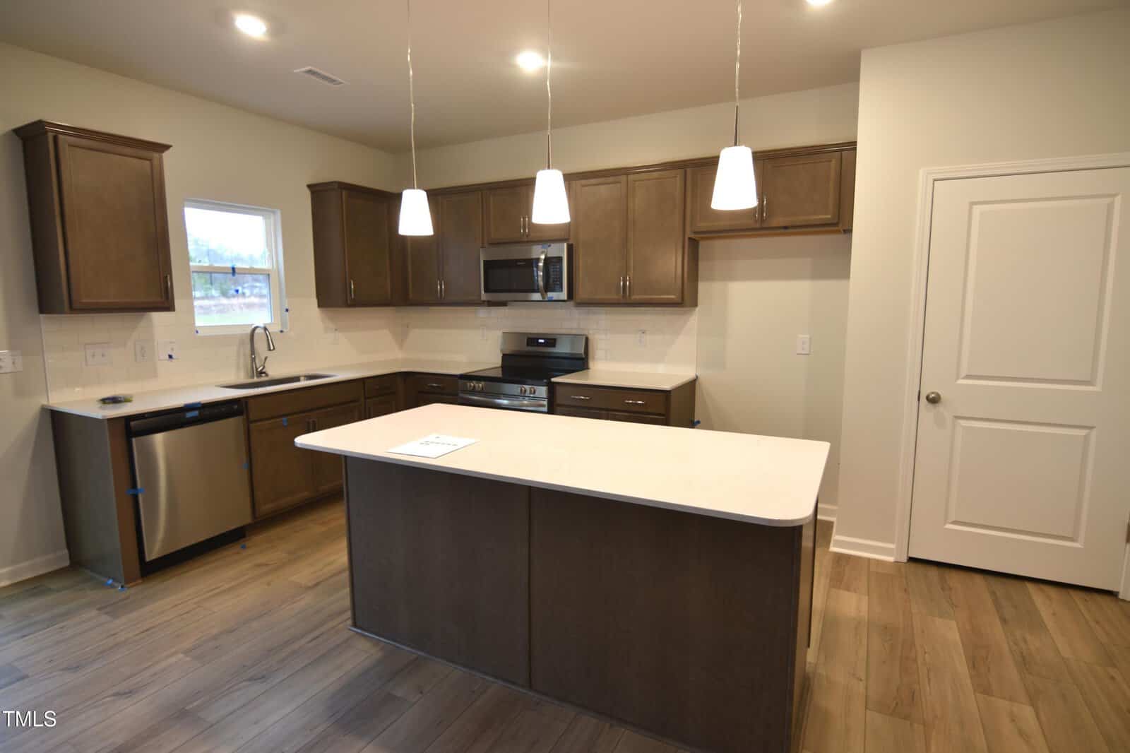 Modern kitchen with brown cabinets, stainless steel appliances, a center island with a white countertop, three pendant lights, wood flooring, and a closed white door on the right.