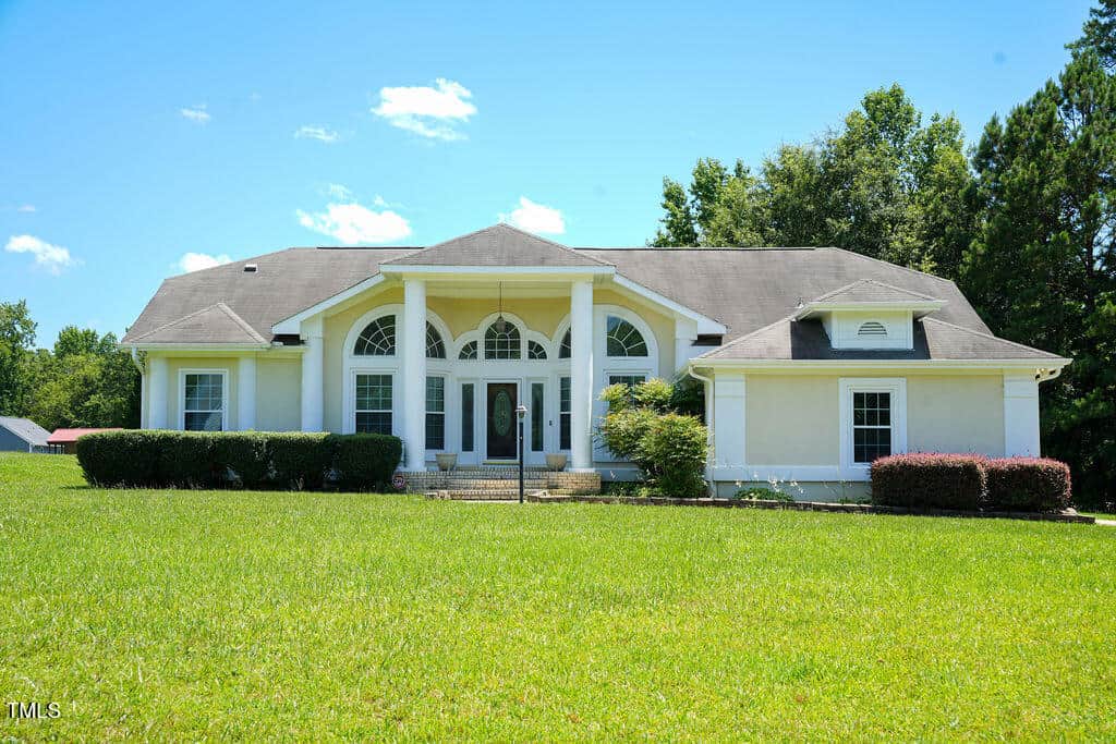 A white, single-story house with columns at the entrance, arched windows, and a shingled roof sits on a large, grassy lawn at 6101 Valentine Street in Zebulon. Shrubs and trees surround the home under a clear blue sky.
