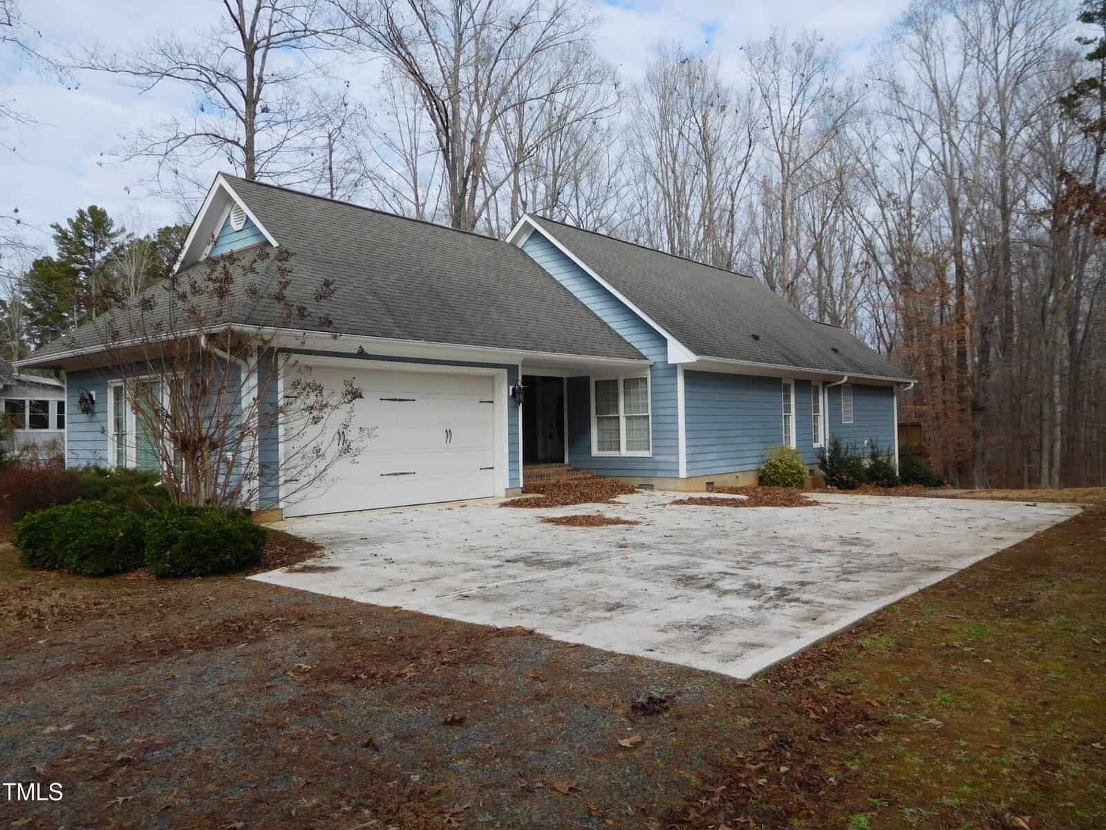 The single-story house at 513 Port Drive, Henderson, features pale blue siding and a gray roof, surrounded by bare trees. It boasts a double garage and a wide driveway. The mostly brown yard hosts some shrubs near the house, set under an overcast sky.