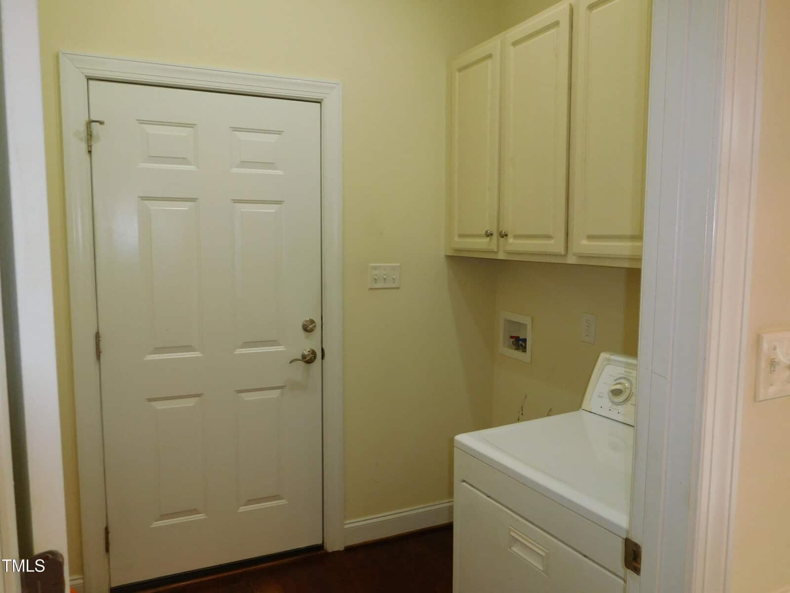 A laundry room with a white washing machine on the right, beige cabinets above, and a closed white door on the left. The walls are painted light yellow, and the floor is dark wood.