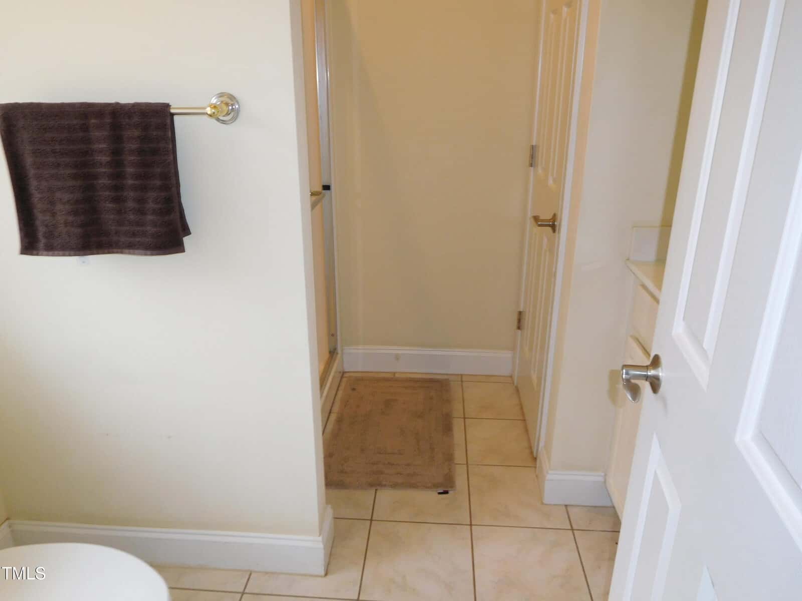 A bathroom with beige walls features a tiled floor and a folded brown towel hanging on a chrome towel rack. A bath mat is placed in front of a shower with a glass door. Two white doors are visible, one partially open.