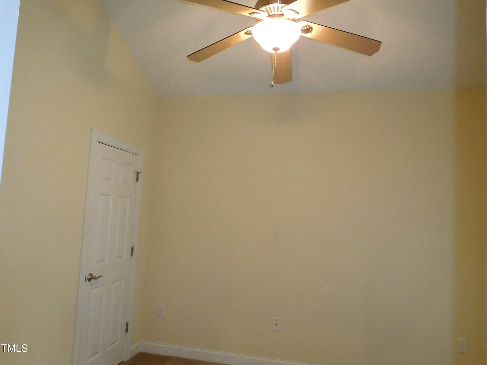A small room with pale yellow walls features a white door and a wooden ceiling fan with a light fixture. The ceiling is sloped, and the room appears unfurnished. Carpet is visible on the floor.