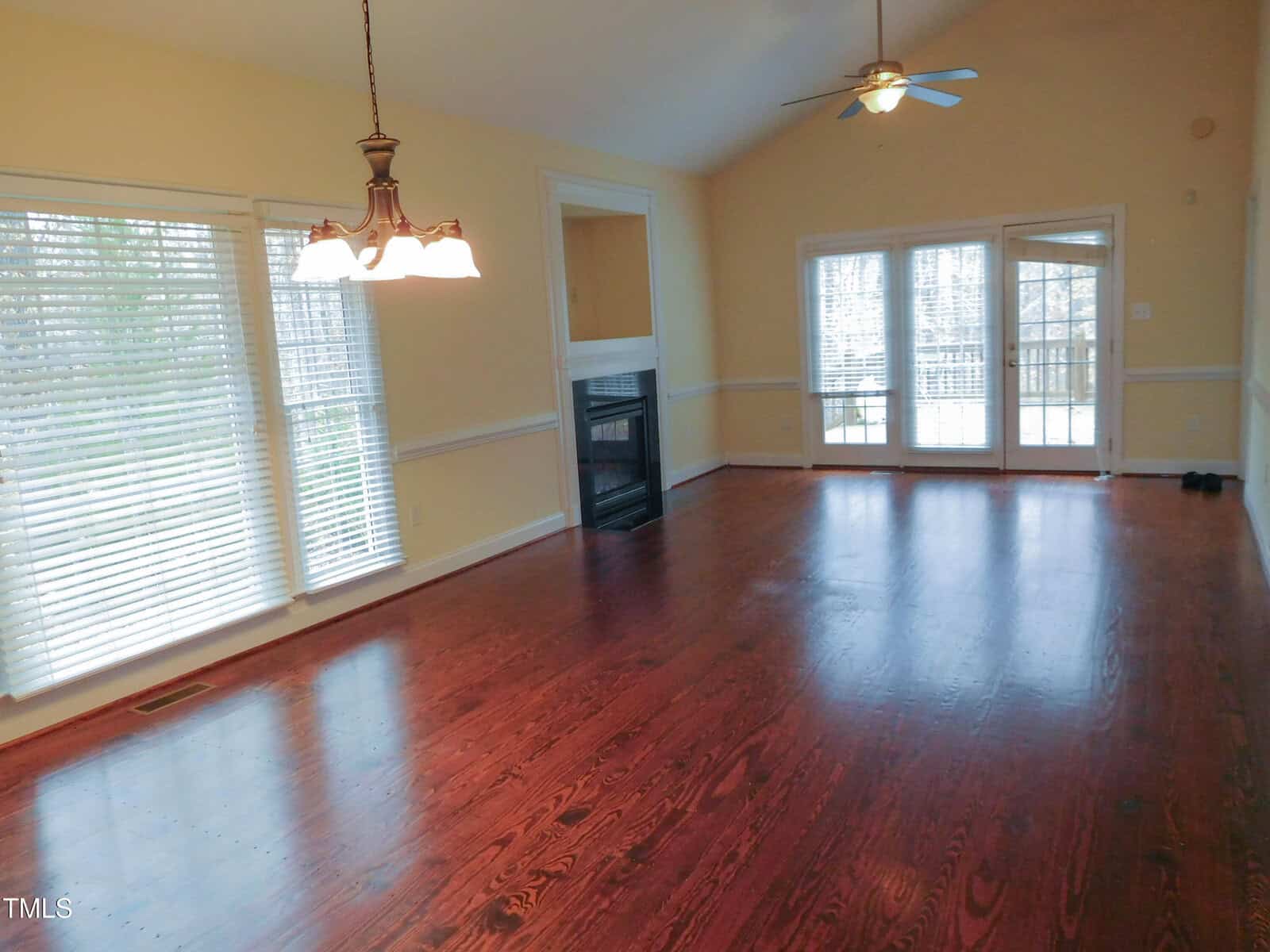 A spacious living room with wooden flooring and cream-colored walls. Large windows with blinds allow natural light. A hanging light fixture is above, and a ceiling fan is on the high ceiling. French doors open onto a terrace.