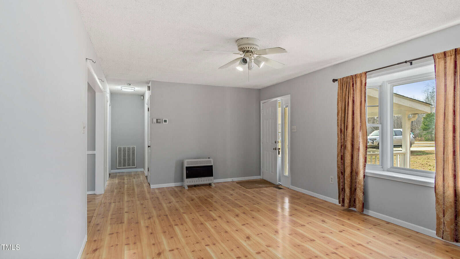 A vacant room with light wood flooring, pale gray walls, and a white ceiling fan. There's a window with red curtains on the right and a visible open door. A wall heater is mounted in the hallway leading to additional rooms.