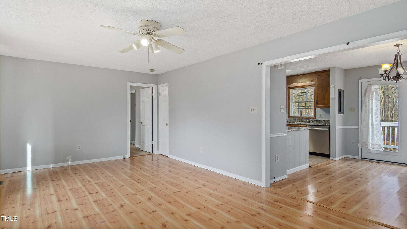 A spacious, empty living room with light wooden floors and gray walls. A ceiling fan is centered on the white ceiling. The room connects to a kitchen with stainless steel appliances and a dining area with a glass door leading outside.