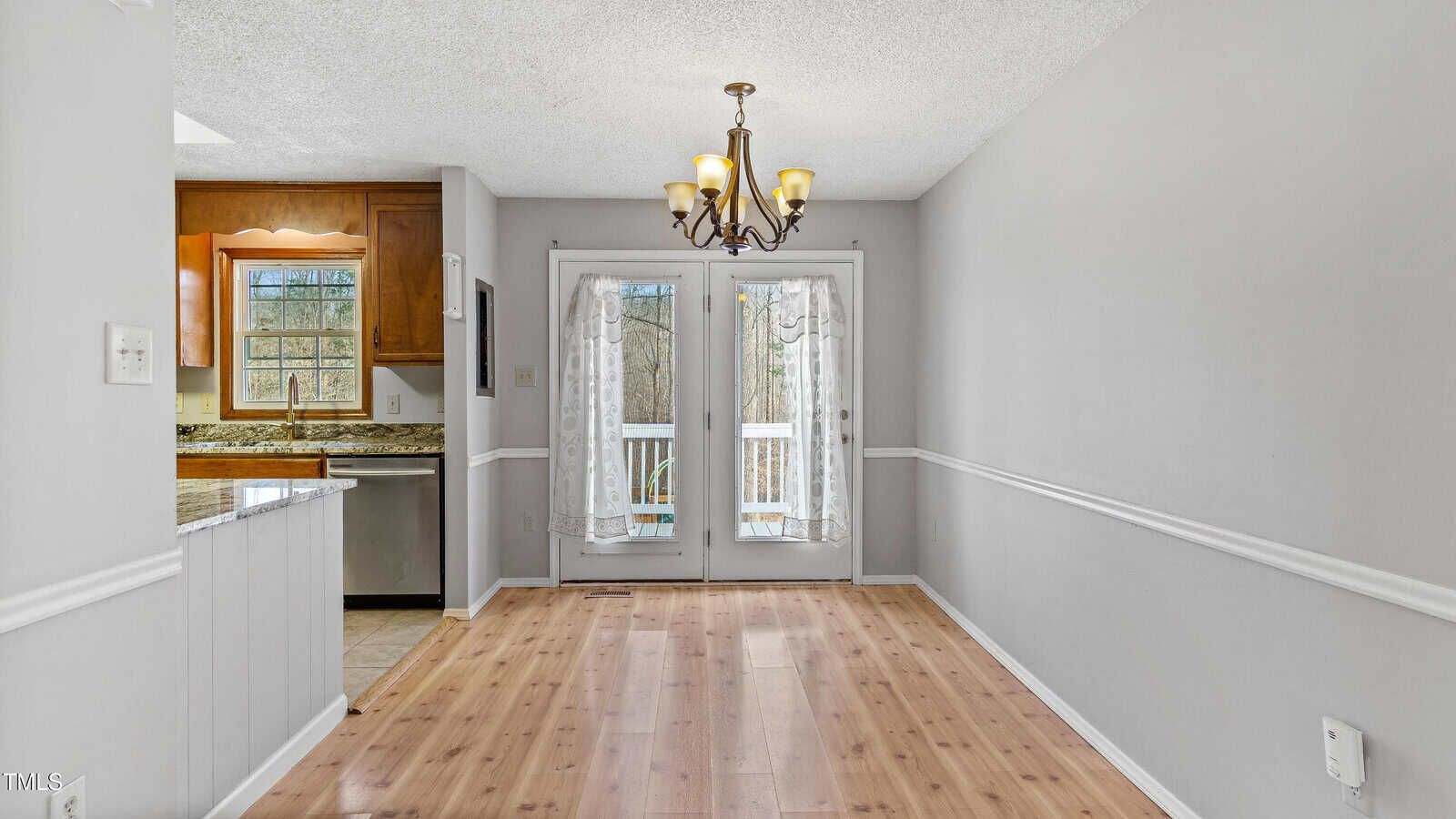 A dining area with light wood flooring and gray walls featuring a central chandelier. Glass double doors with sheer curtains lead to an outdoor deck. A compact kitchen with wooden cabinets and a granite countertop is visible to the left.