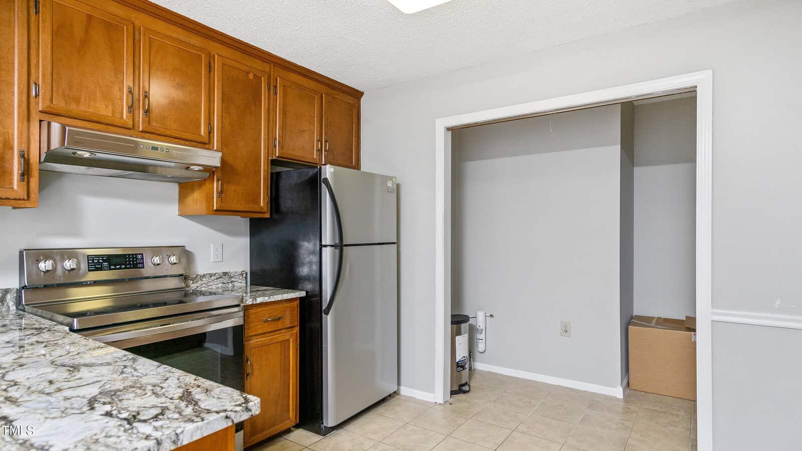 A kitchen with wooden cabinets and a stainless steel oven and refrigerator. The countertop is marble-patterned, and the floor is tiled. An open closet with a white door frame is visible to the right with a box and a lamp stand partially inside.