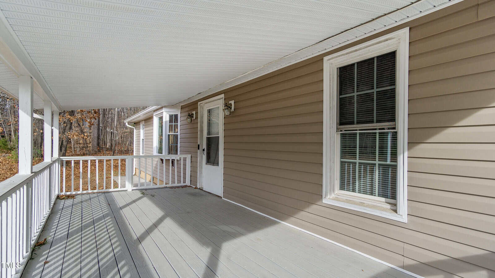 A wooden porch with a light gray floor and white railings attached to a house with beige siding. Two windows with blinds and a white door with outdoor lamps are on the house. The porch overlooks a wooded area.