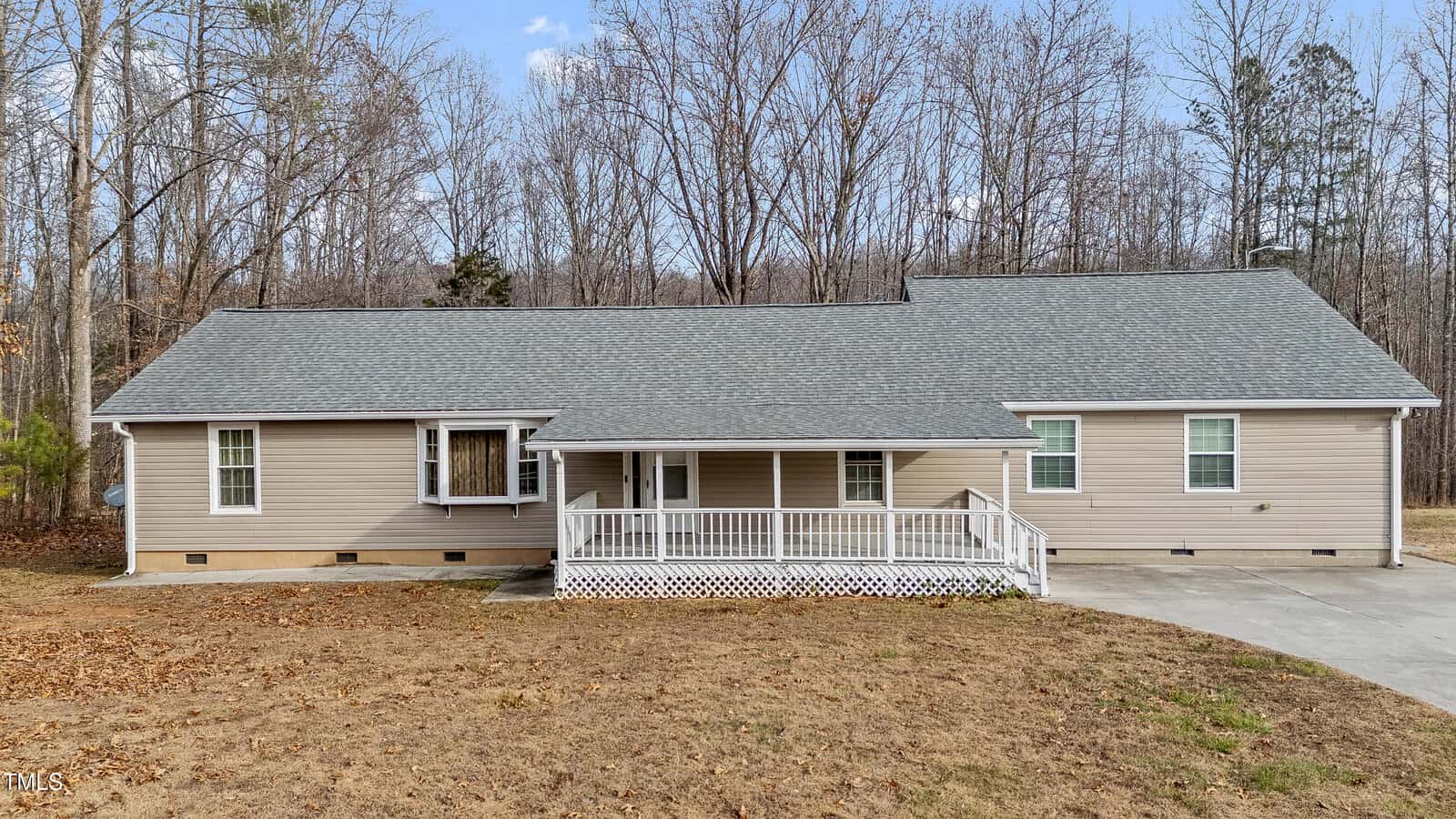 A single-story beige house with a gray roof sits amid a wooded area. It features a white railing on the front porch and a concrete driveway leading to the entrance, surrounded by bare trees and a grassy yard.