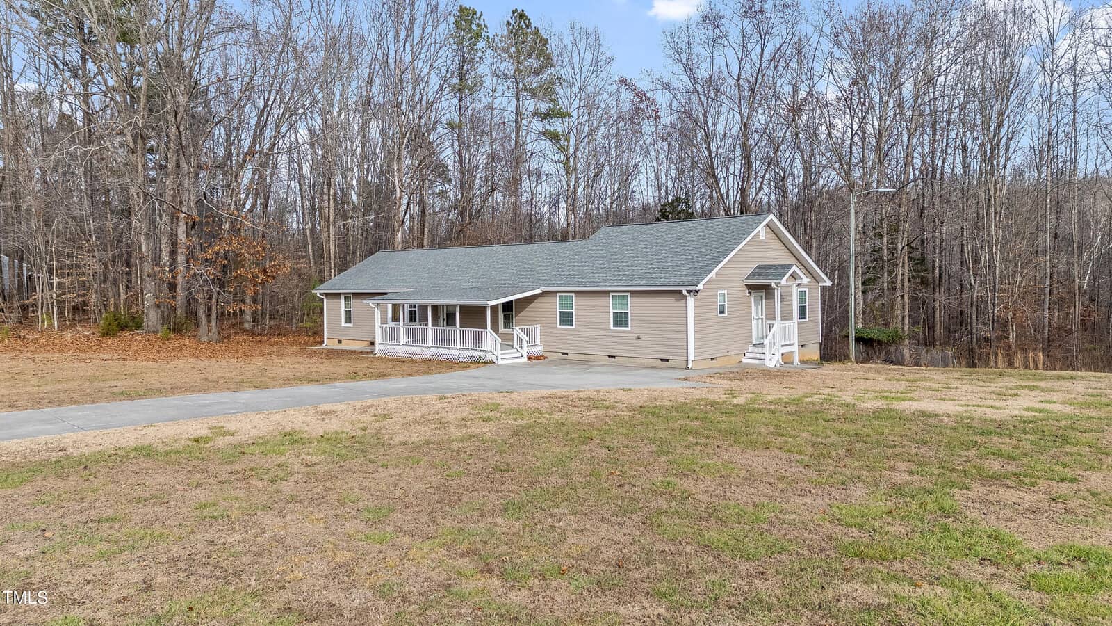 A single-story house with gray siding and a green roof is surrounded by bare trees. The home features a large front porch with white railings and a ramp. A concrete driveway leads up to the house, and the lawn is mostly brown grass.