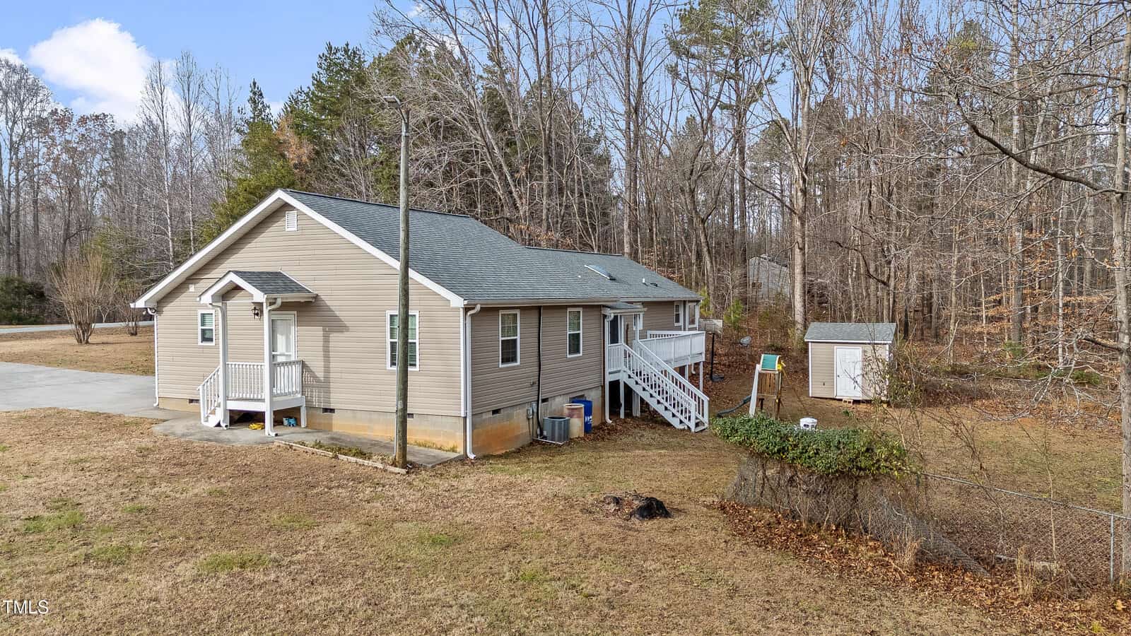 A single-story beige house with a gray roof is surrounded by a leafless forest. A small shed and a fenced area are in the backyard. A concrete driveway leads to the front steps. The sky is clear and sunny.