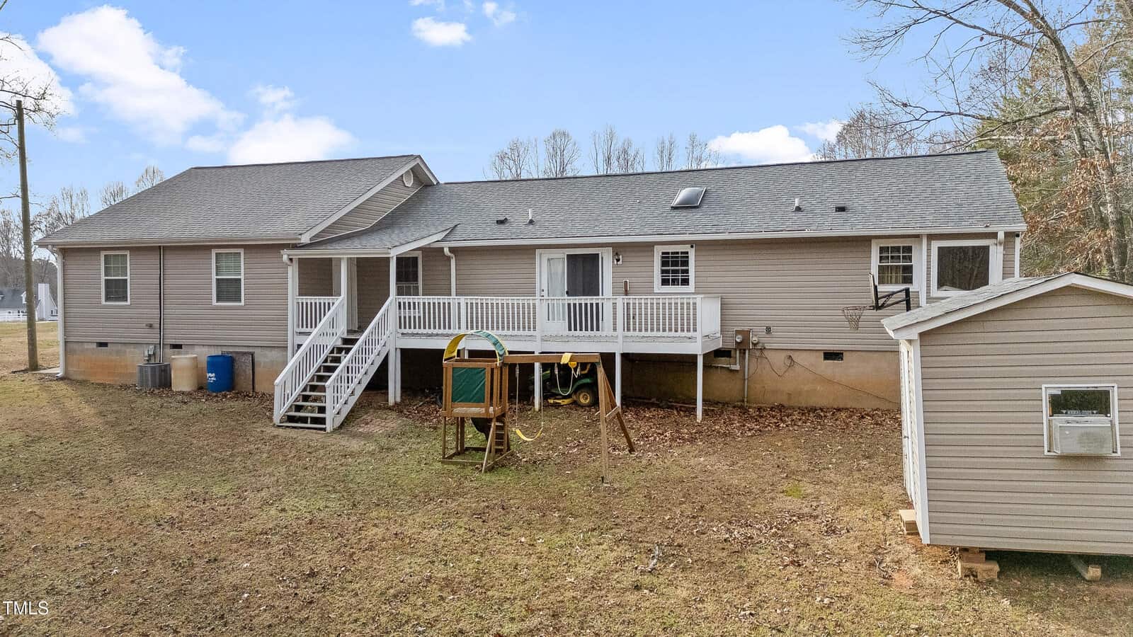 A single-story gray house with a white railing around the back deck. A set of stairs leads to the grassy yard, where there is a small wooden playset. A shed is visible on the right. Bare trees are in the background under a cloudy sky.