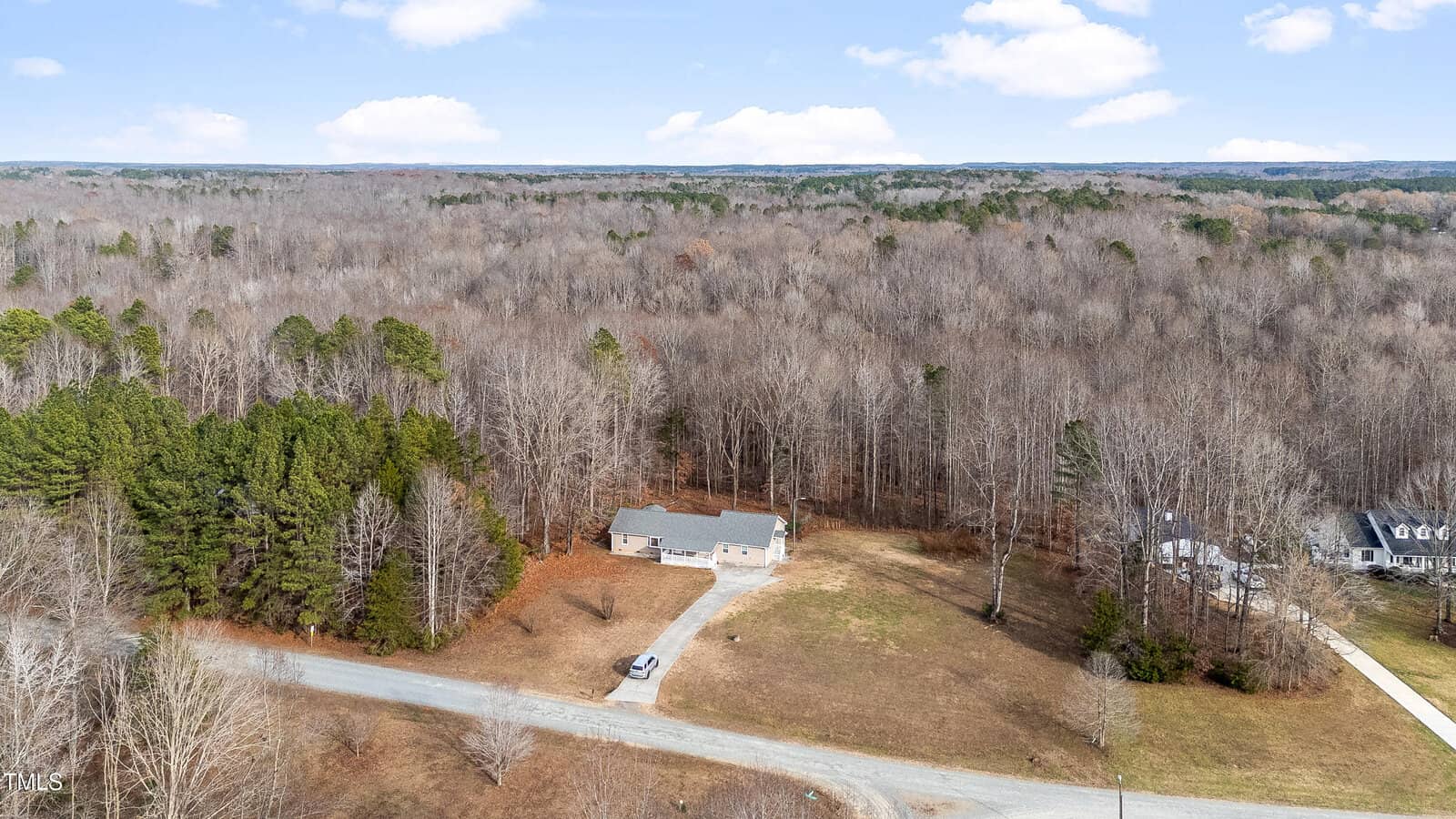 Aerial view of the rural landscape at 5584 Wingate Creek Road in Oxford, showcasing a single house nestled amid dense woodlands. The property features a large grassy area in front, a driveway, and an adjacent road. Another house is partly visible to the right, with sparse trees stretching into the distance.