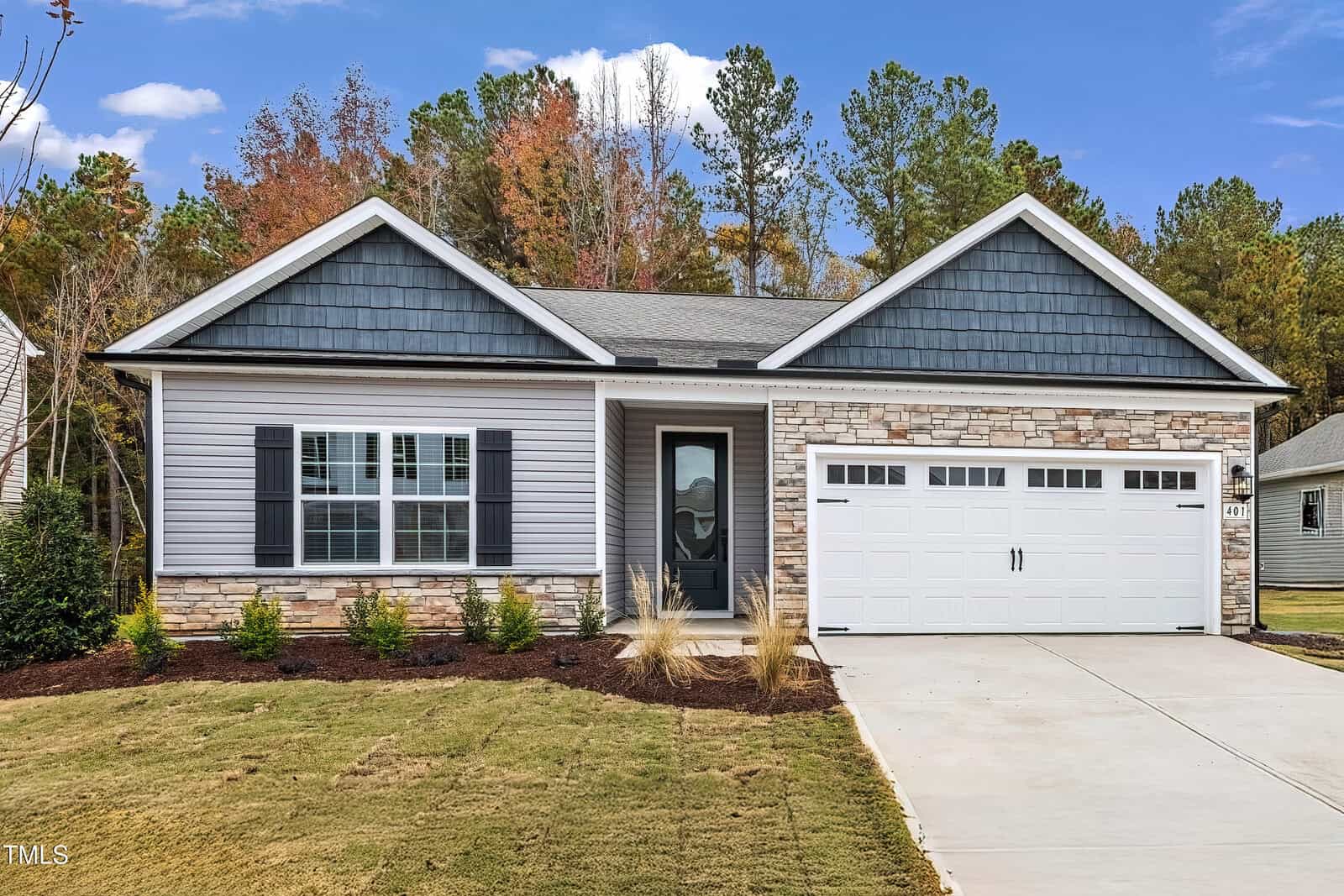 Single-story suburban house with gray siding, stone accents, black shutters, and a two-car white garage. There is a front door with a window and a small front yard with grass and shrubs. Trees and blue sky are in the background.