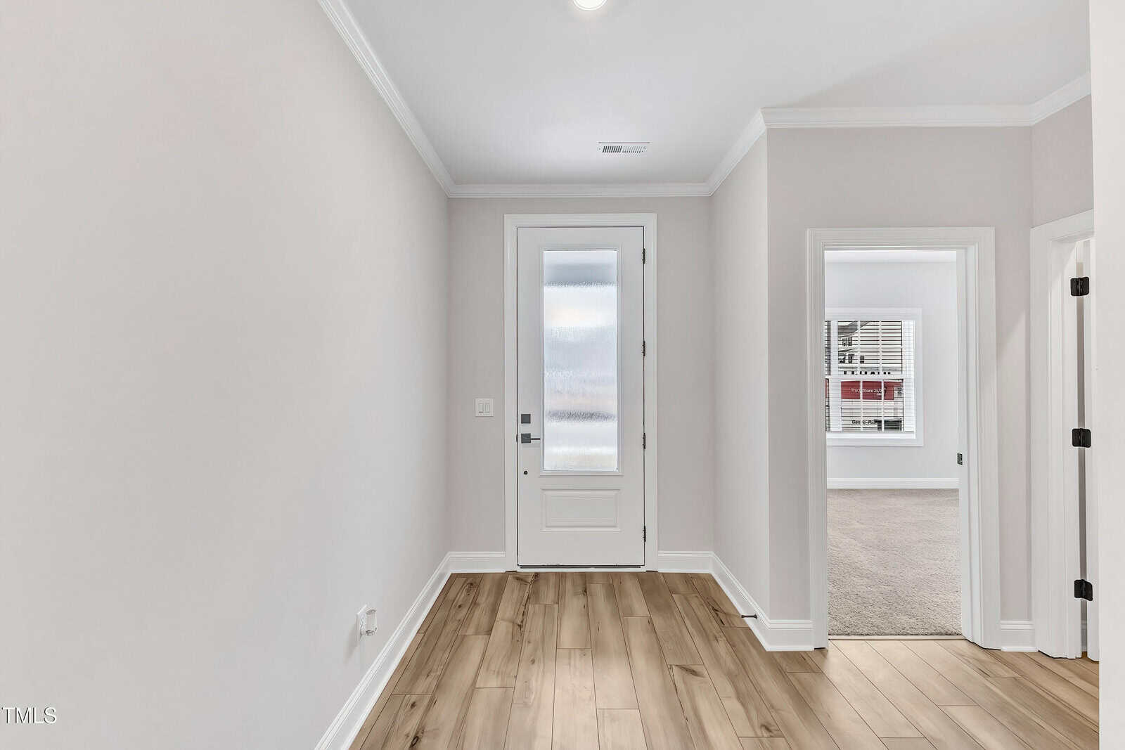 A bright entryway with light-colored walls, wooden floor, and a white door with frosted glass. Two open doorways lead to carpeted rooms, and white trim accents the space.